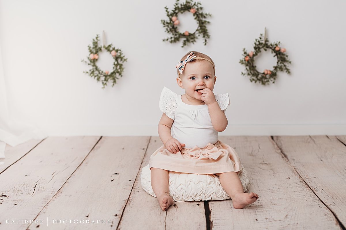 Baby in white outfit sitting on wooden floor with delicate wreaths on wall.