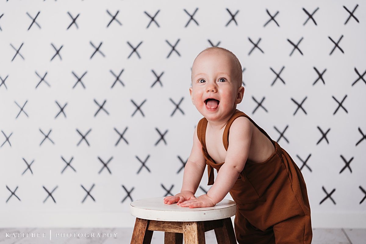 Baby in brown outfit stands at small wooden stool against patterned white backdrop.