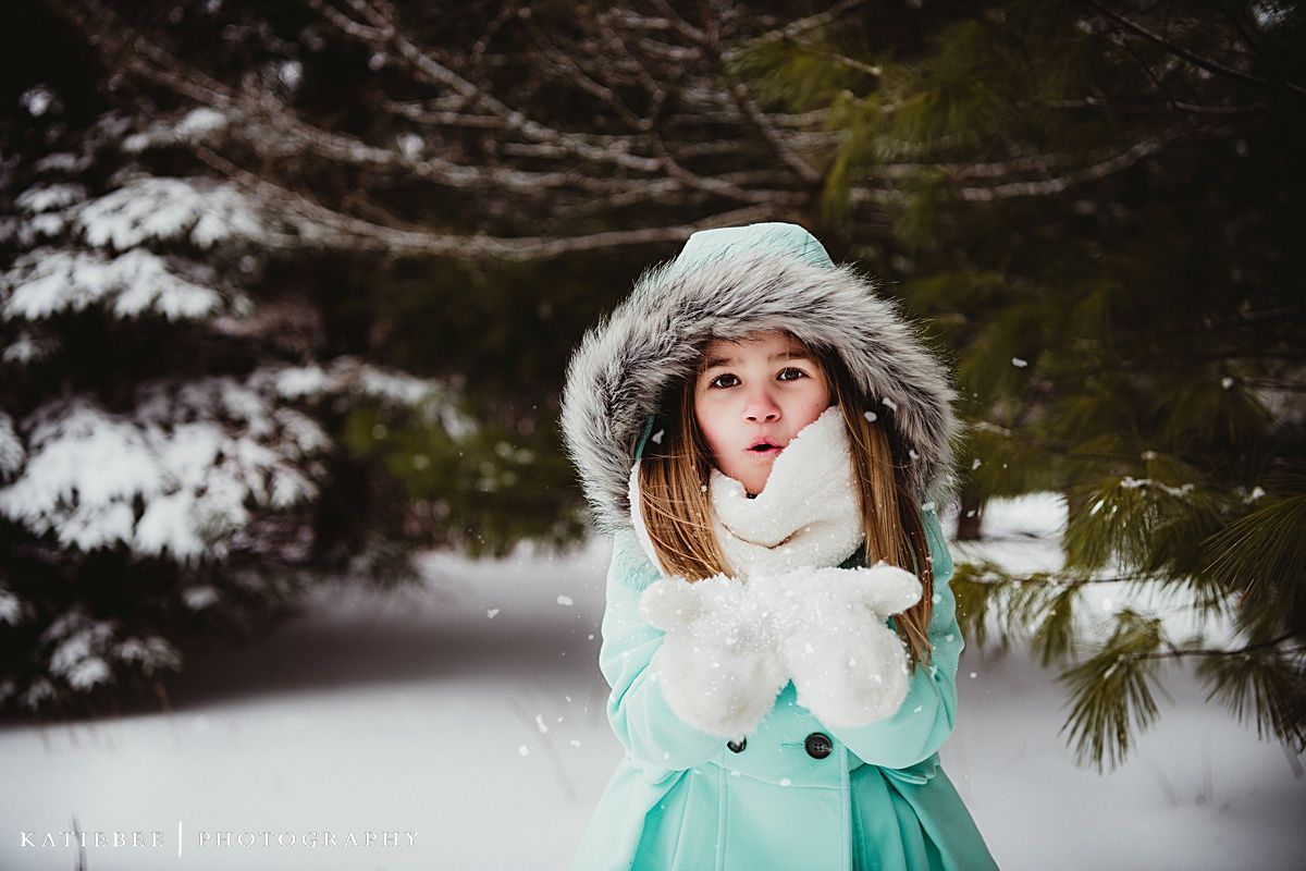 A winter portrait showing a mint green coat and fur-trimmed hood against a snowy evergreen forest backdrop.