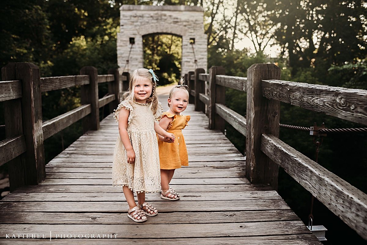Two young children standing on a wooden bridge near a stone archway wearing light colored dresses.