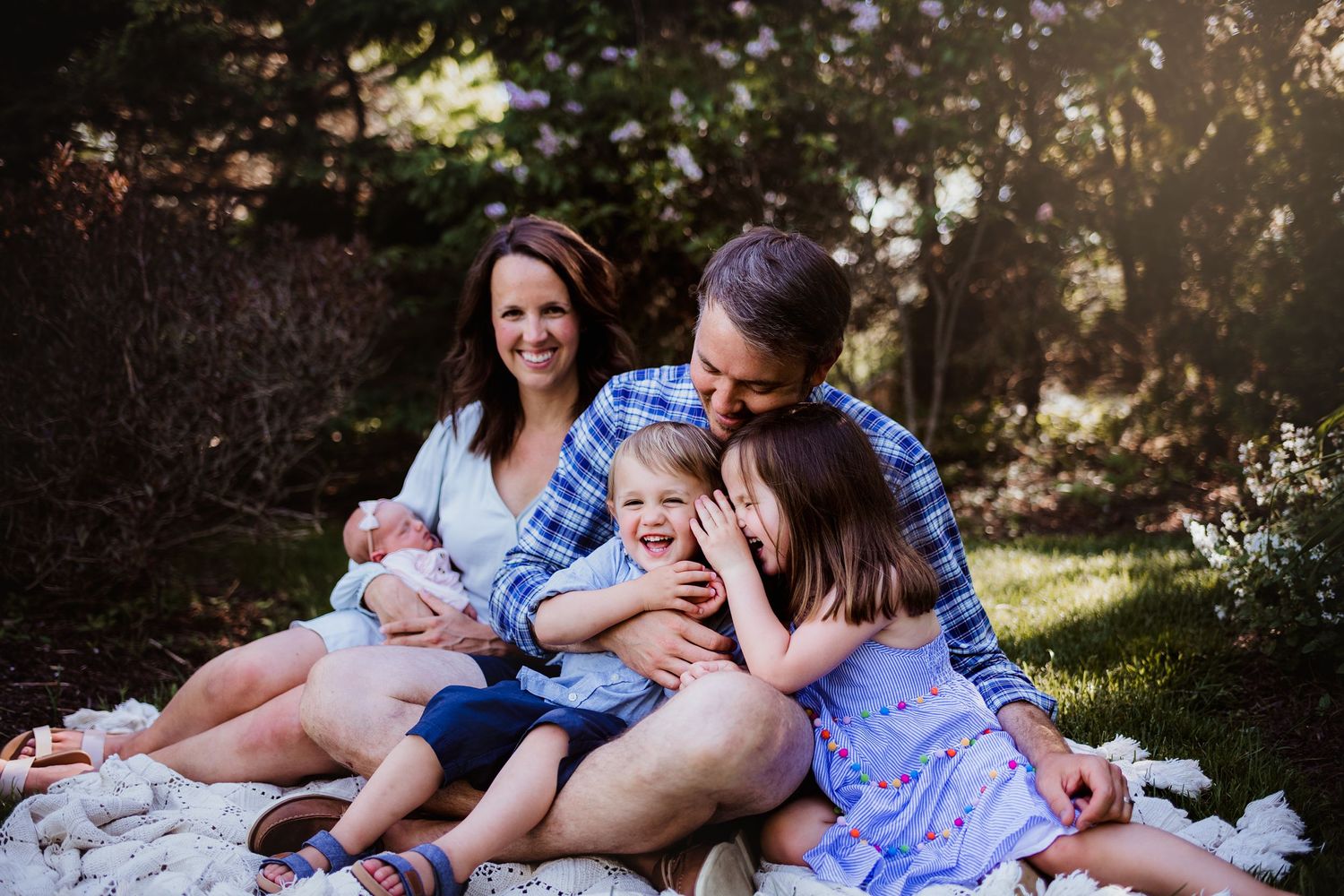 A family cuddles together on a picnic blanket in dappled sunlight.