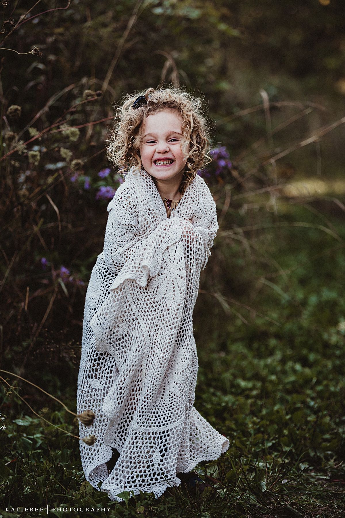 A child wrapped in a white lace shawl laughs while standing in a natural setting.