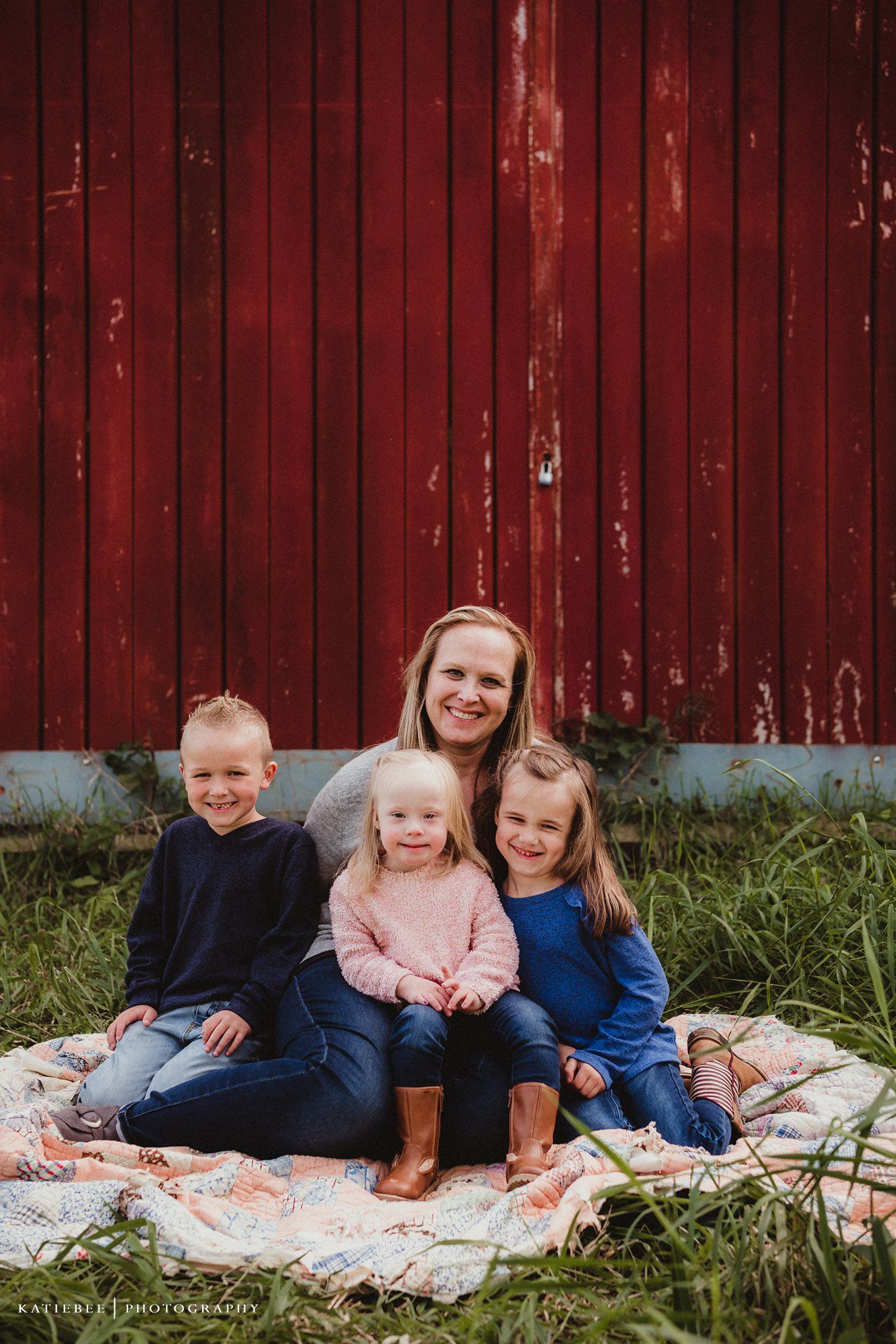 Family group sitting together on a floral blanket against red barn backdrop.