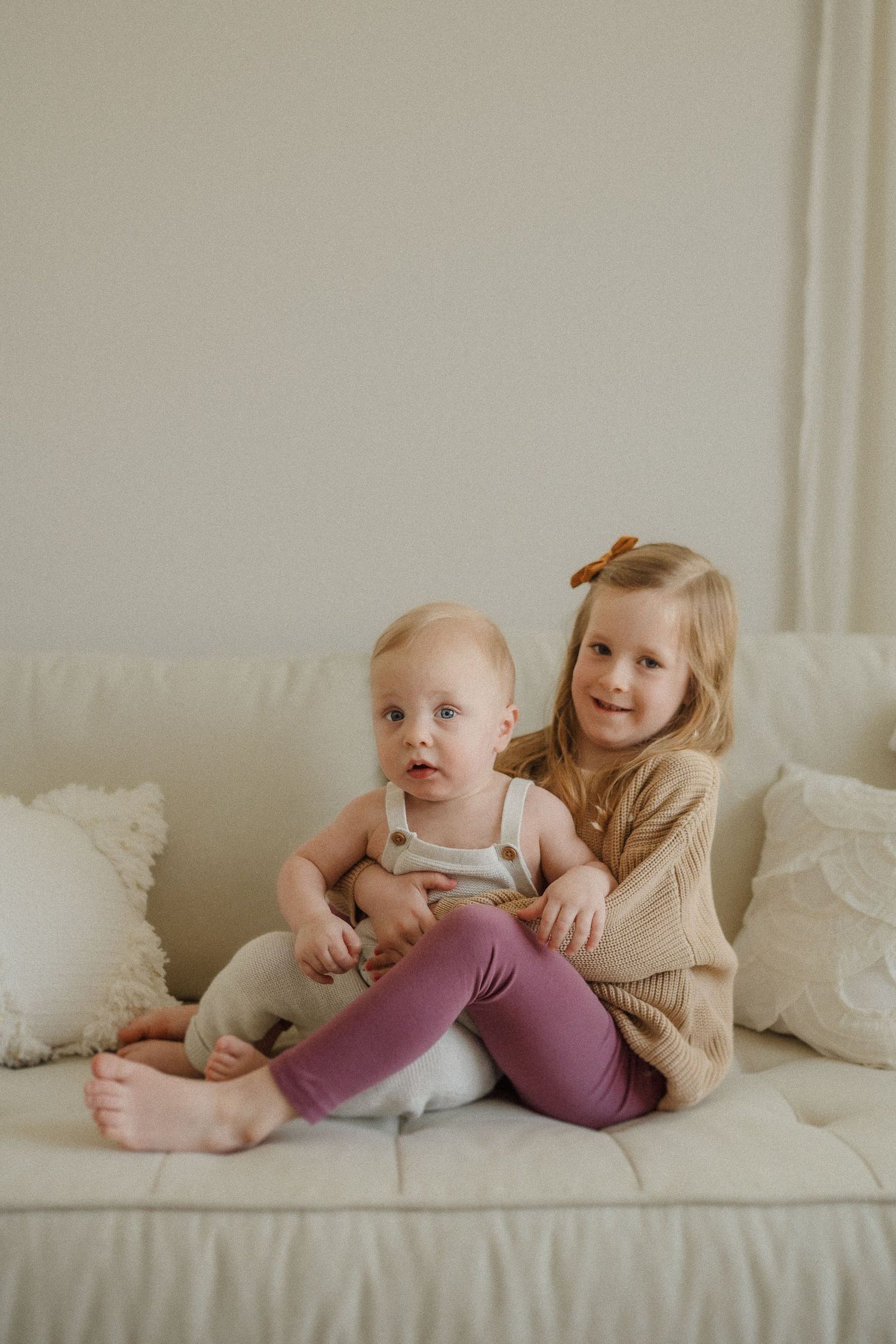 Two children sit together on a cream colored couch with decorative pillows.