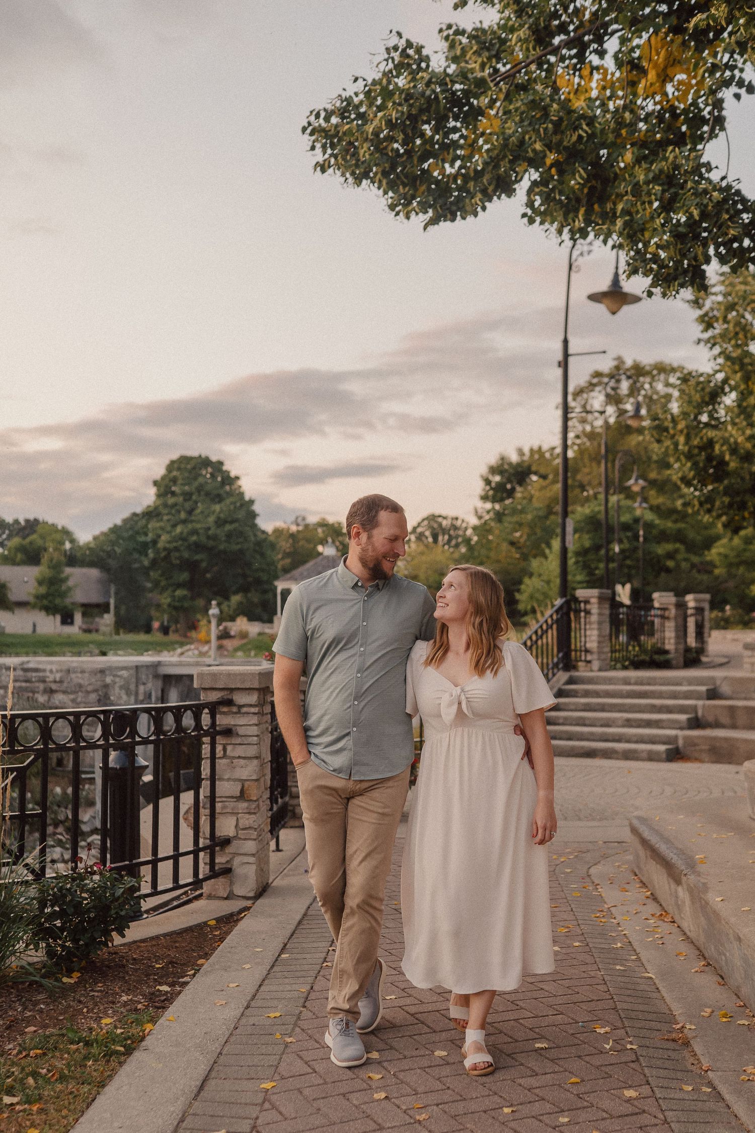 Couple walking together on a paved path at sunset near ornate metal railings.