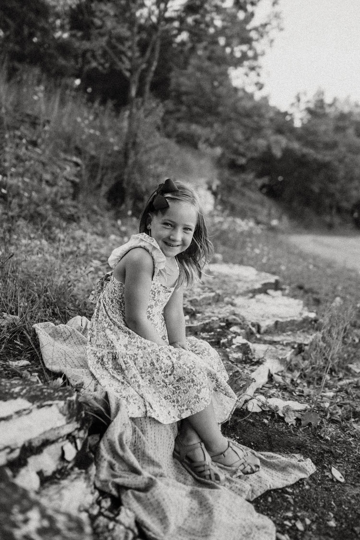 A black and white image shows a child sitting cheerfully on a blanket outdoors.