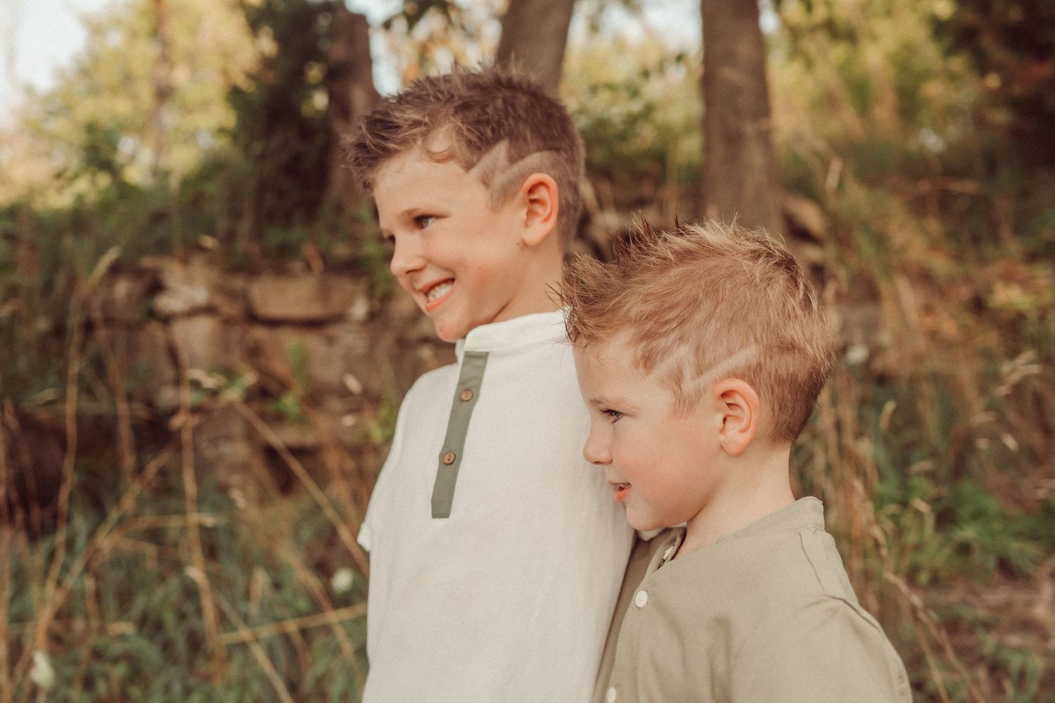 Two children in light colored shirts stand together in an outdoor woodland setting at sunset.