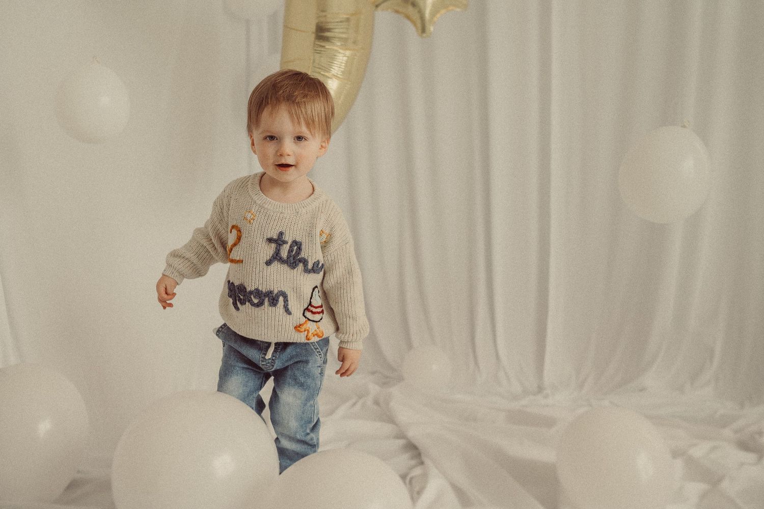 Toddler in a beige sweater and jeans jumping playfully on white bedding with gold star decorations.