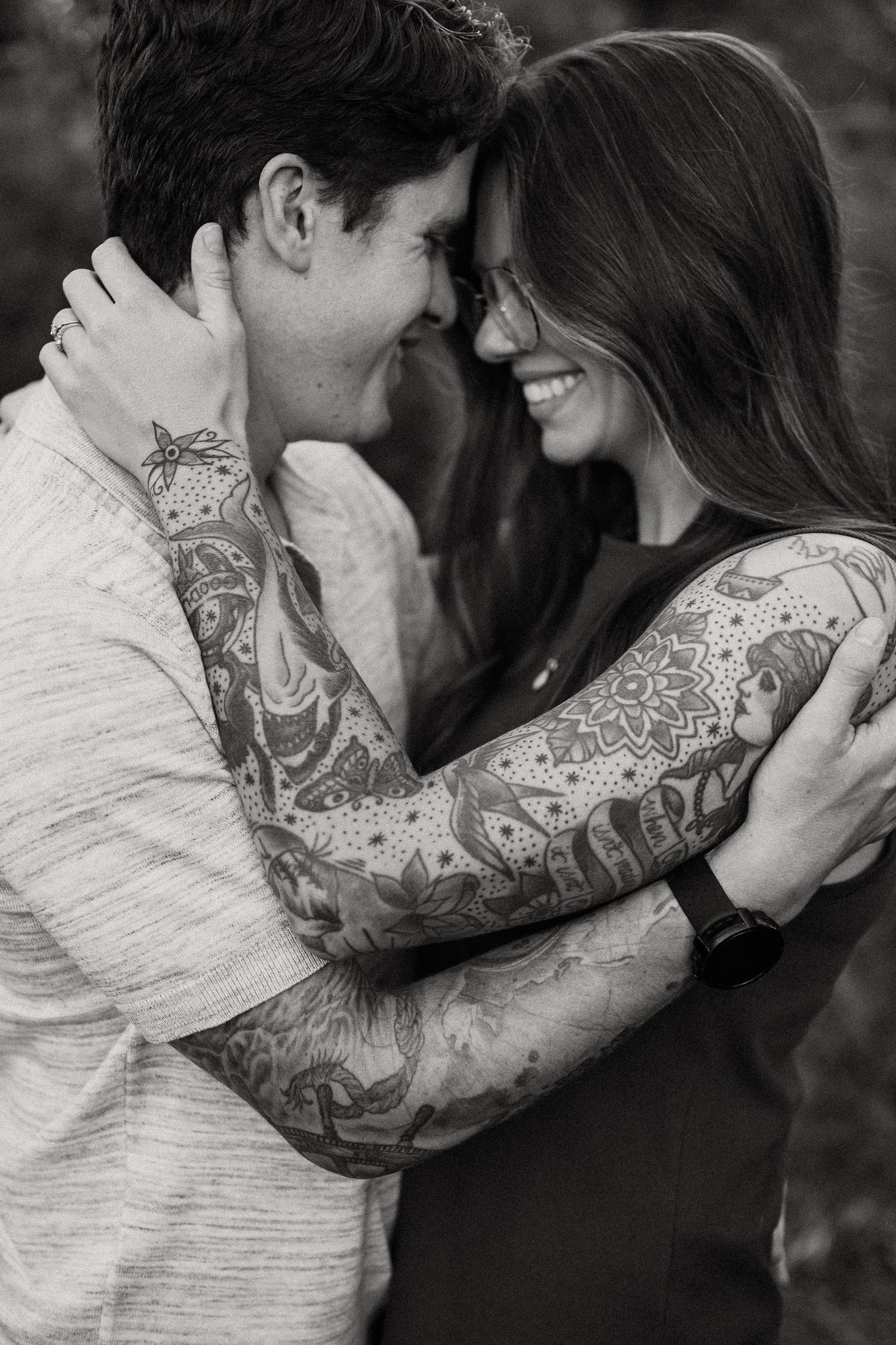 Intimate black and white portrait showing a couple embracing, with one person displaying detailed sleeve tattoos.
