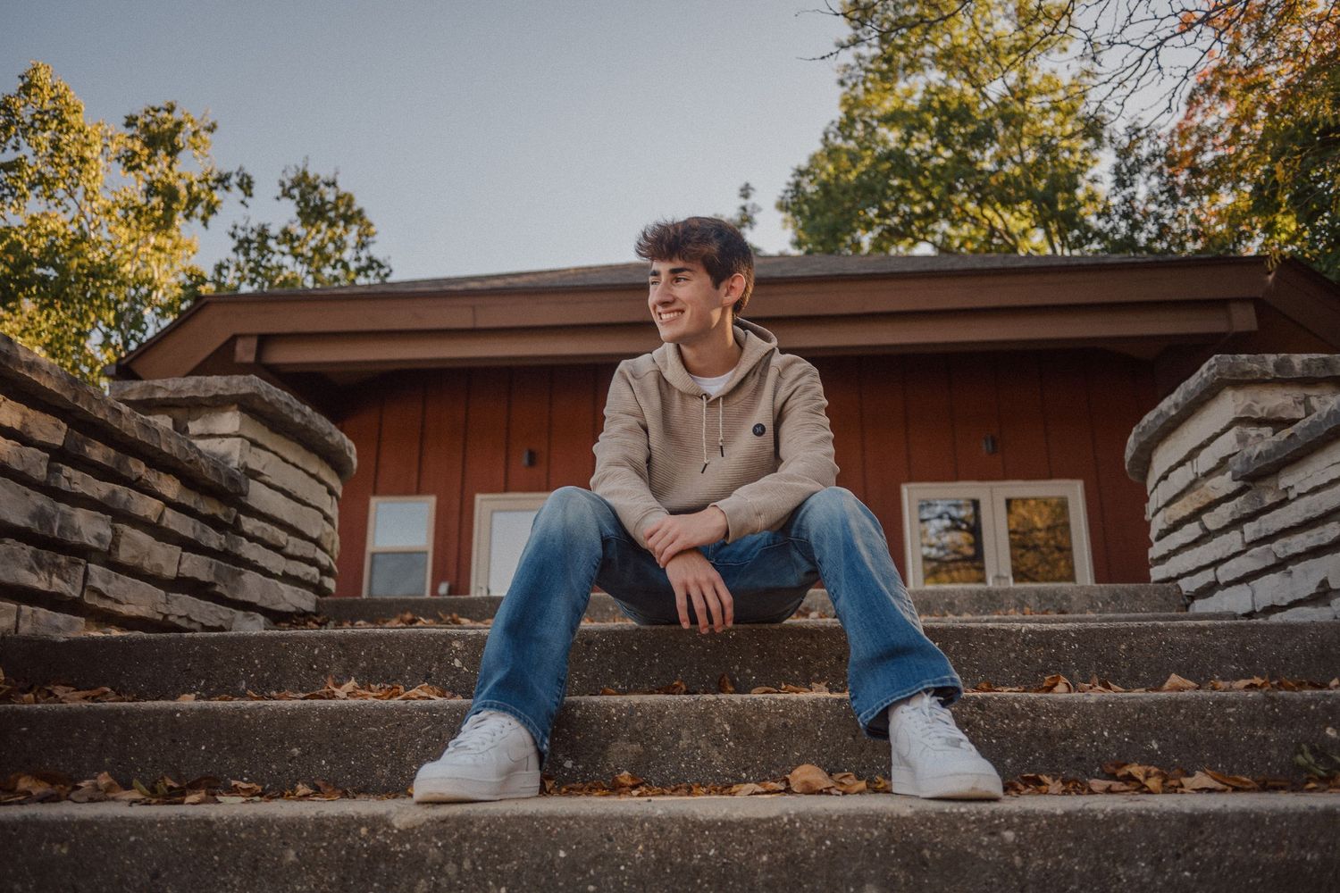 Someone sits on concrete steps in front of a wooden building wearing a hoodie and jeans on an autumn day.