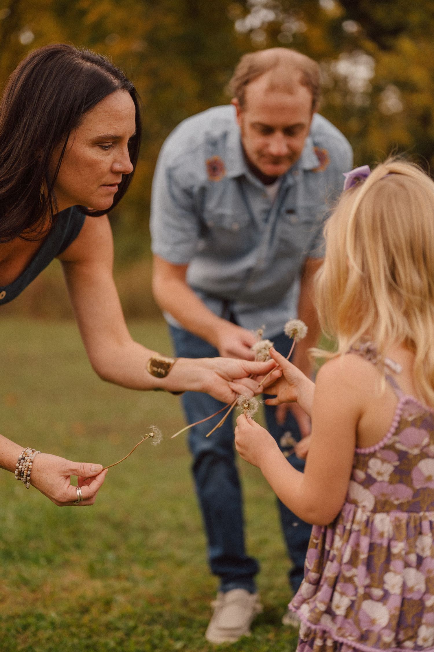 A family interacts playfully on the grass during a fall photoshoot with autumn trees in the background.