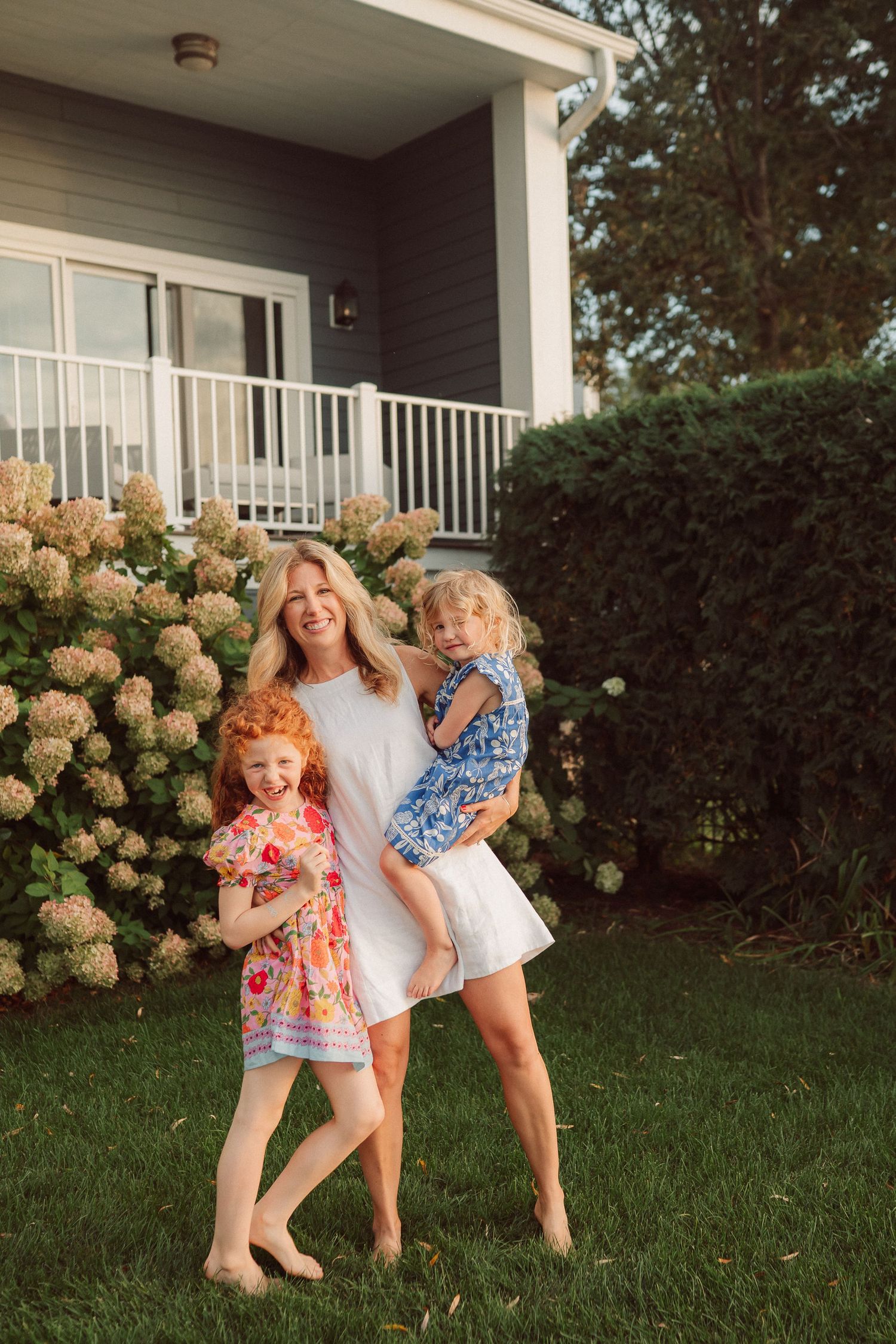 Three children playing and laughing together on a lawn with blooming hydrangea bushes and a white porch in the background.