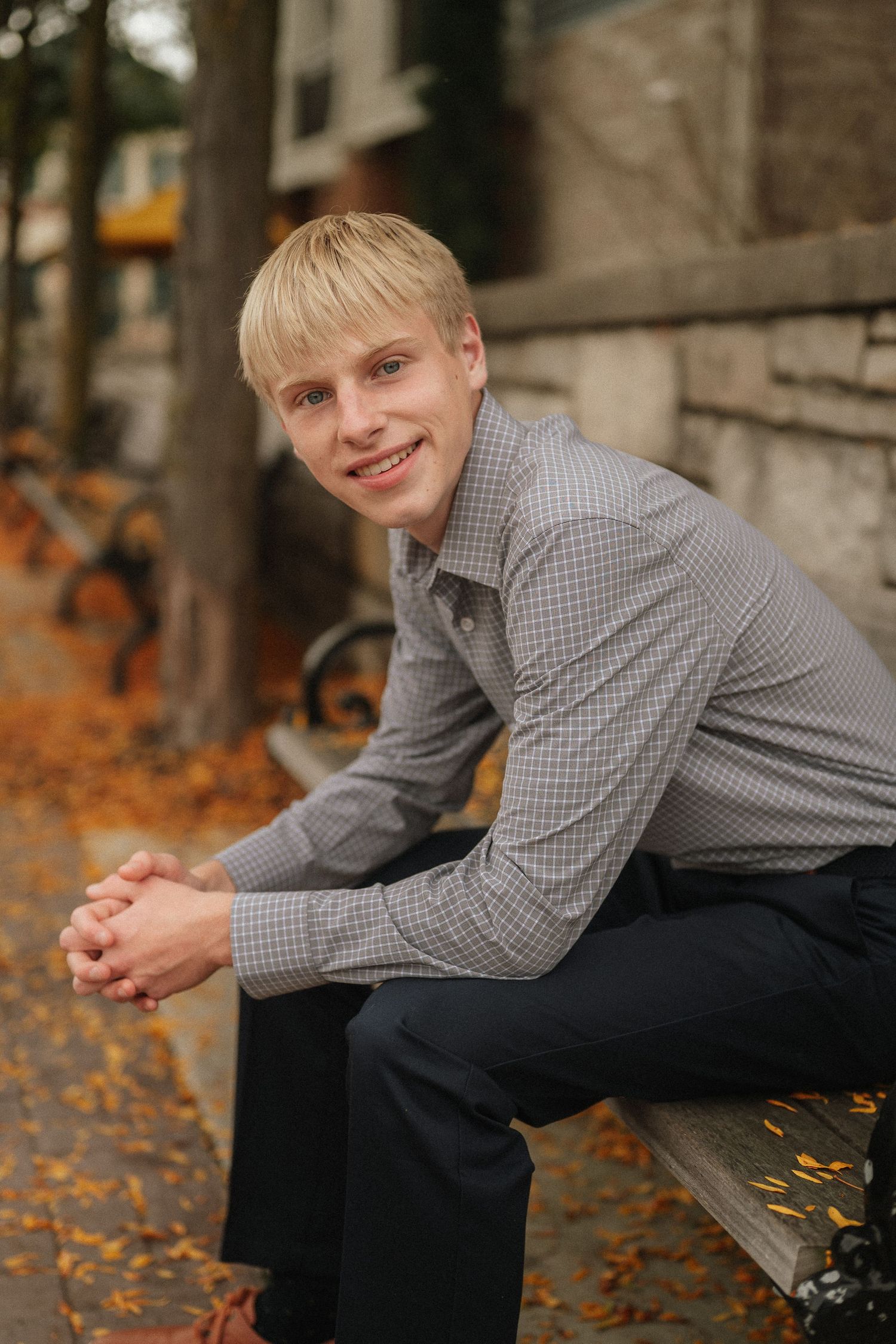A person in a gray checkered shirt and dark pants sits on a bench surrounded by autumn leaves.
