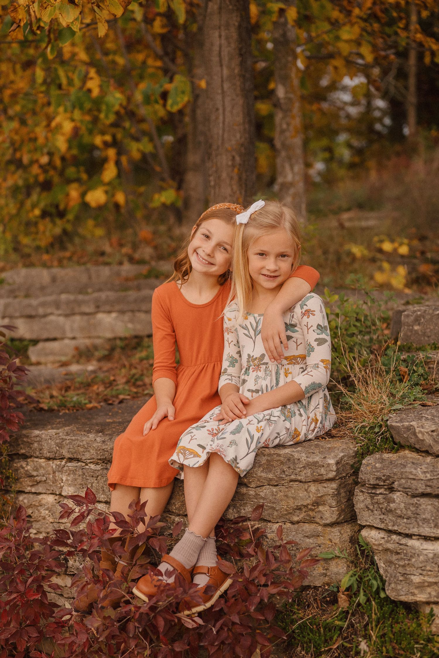 Two children sit together on stone steps surrounded by autumn foliage in a rustic outdoor setting.