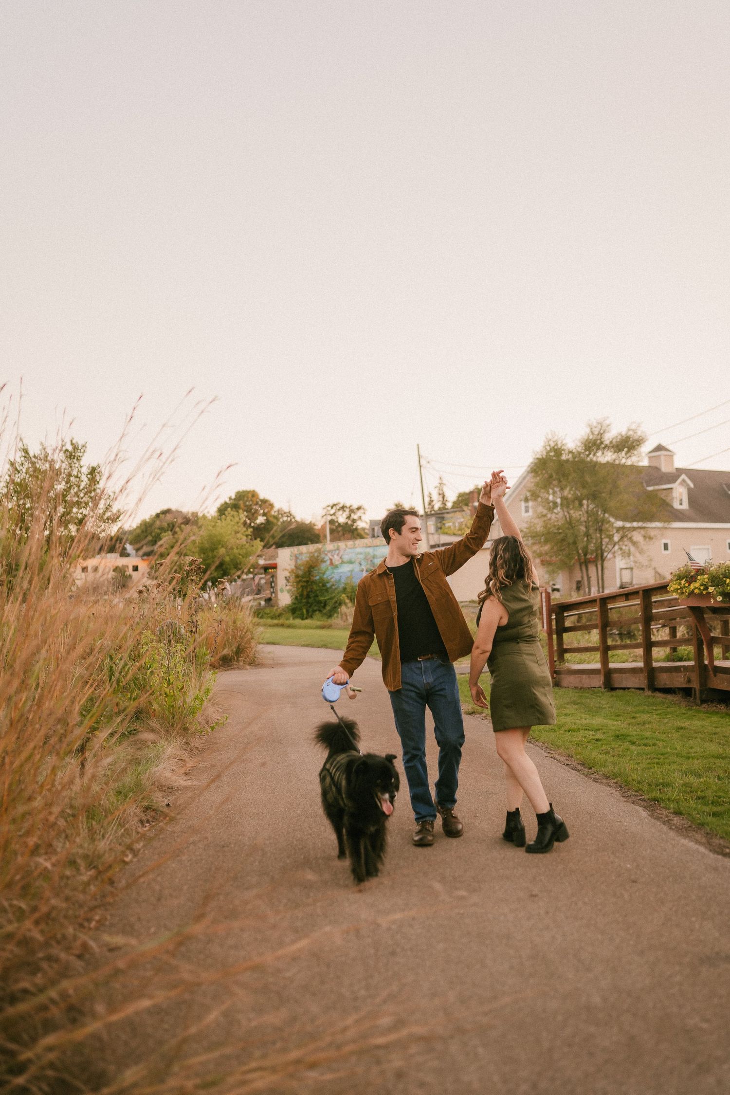 A couple walks their black dog on a path during sunset with rural buildings in background.