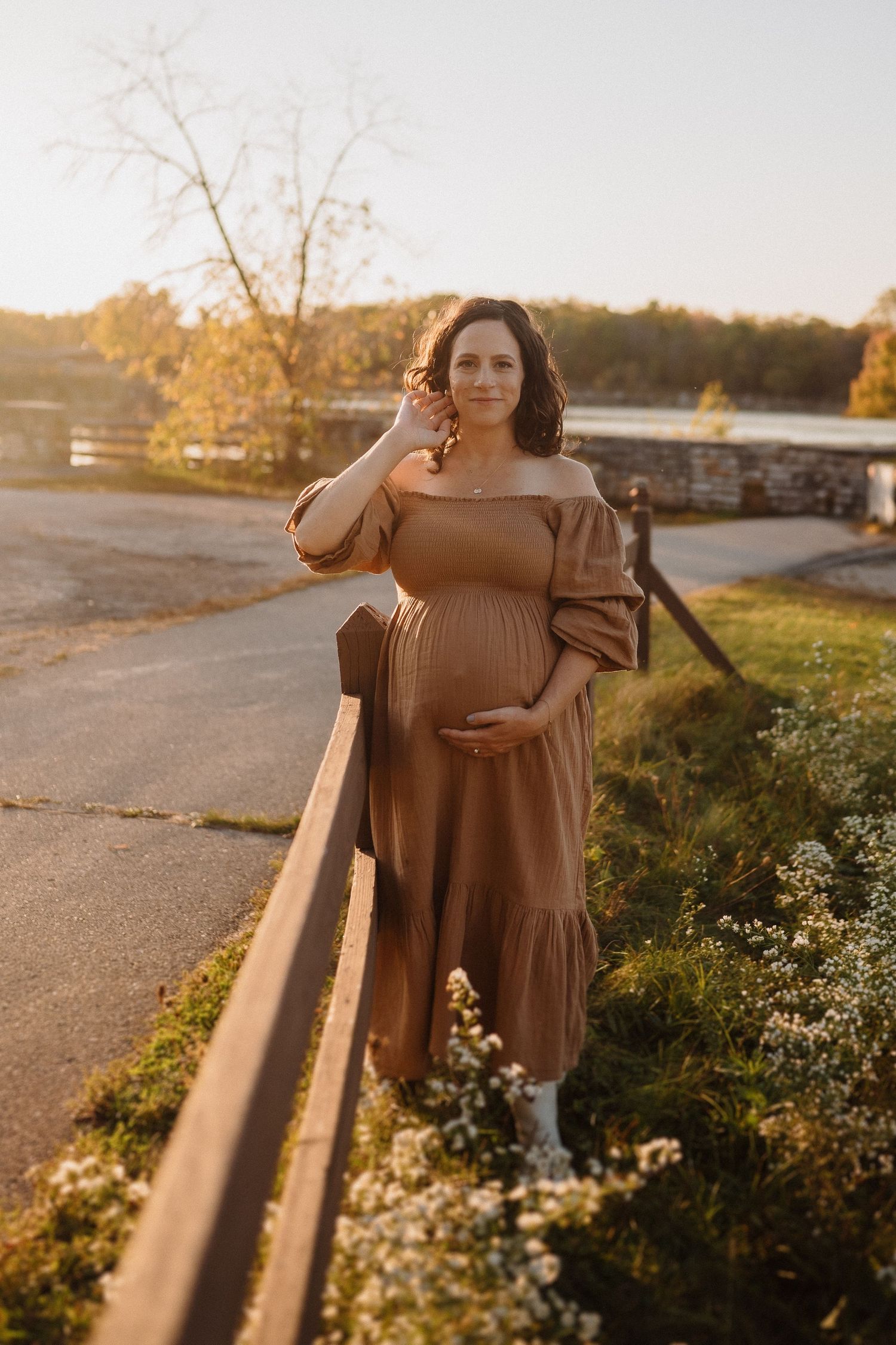 Expectant mother in flowing brown dress poses by fence during golden hour.