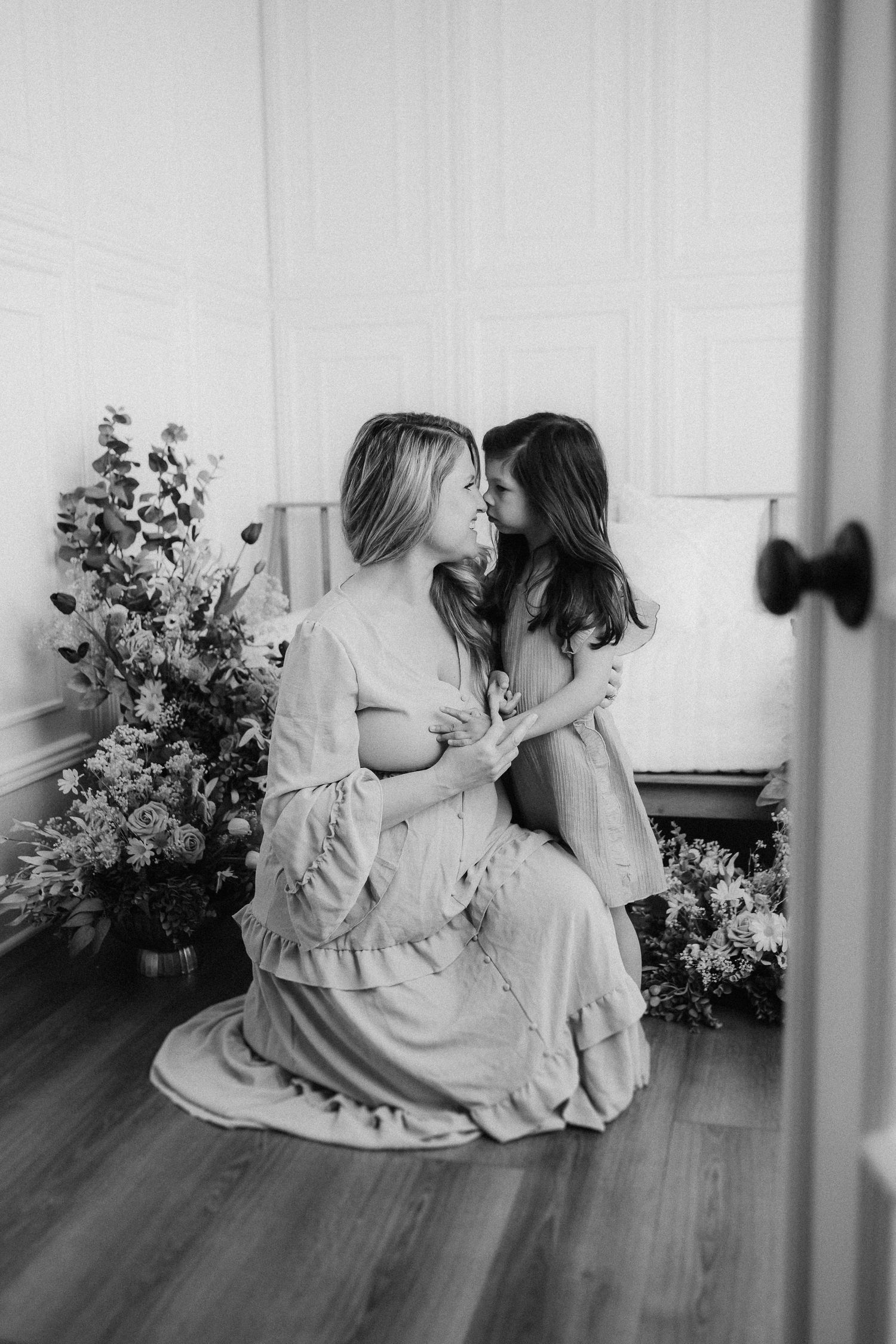 A tender black and white portrait of two people sitting together indoors with floral arrangements on a hardwood floor.