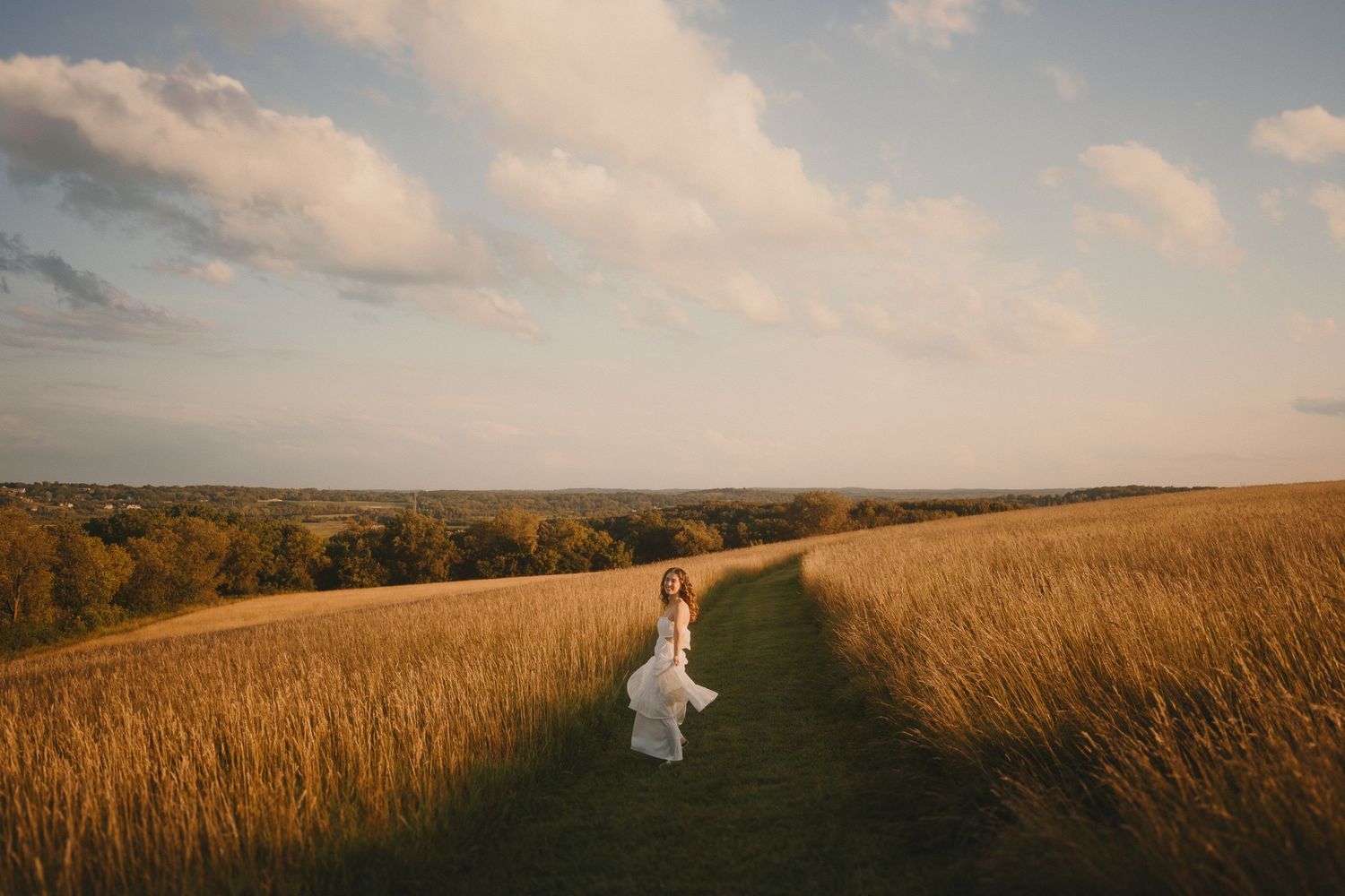 A white dress-clad figure stands alone on a winding path through tall golden grass at sunset.