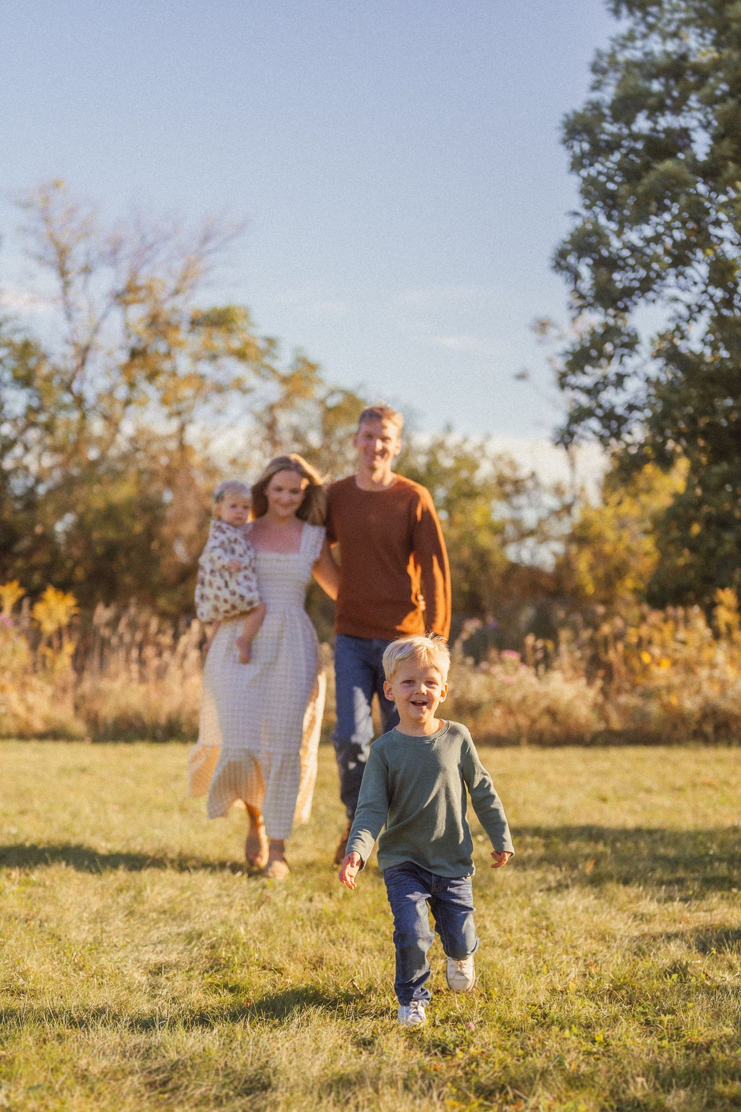 Family walking together in a sunny field at golden hour during autumn with tall grass and trees in background.