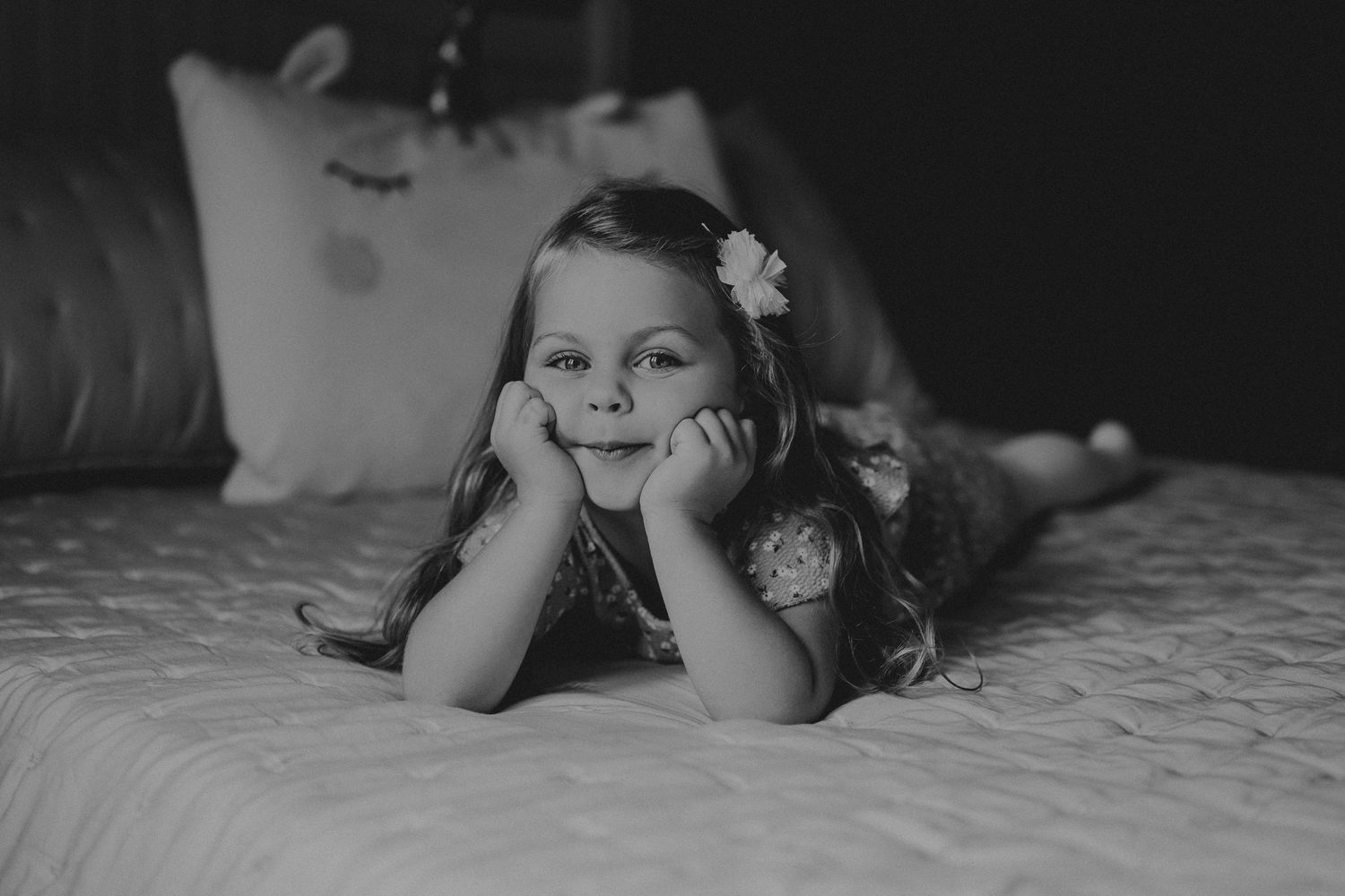 Black and white portrait of child lying on quilted bedspread with flower hair accessory.
