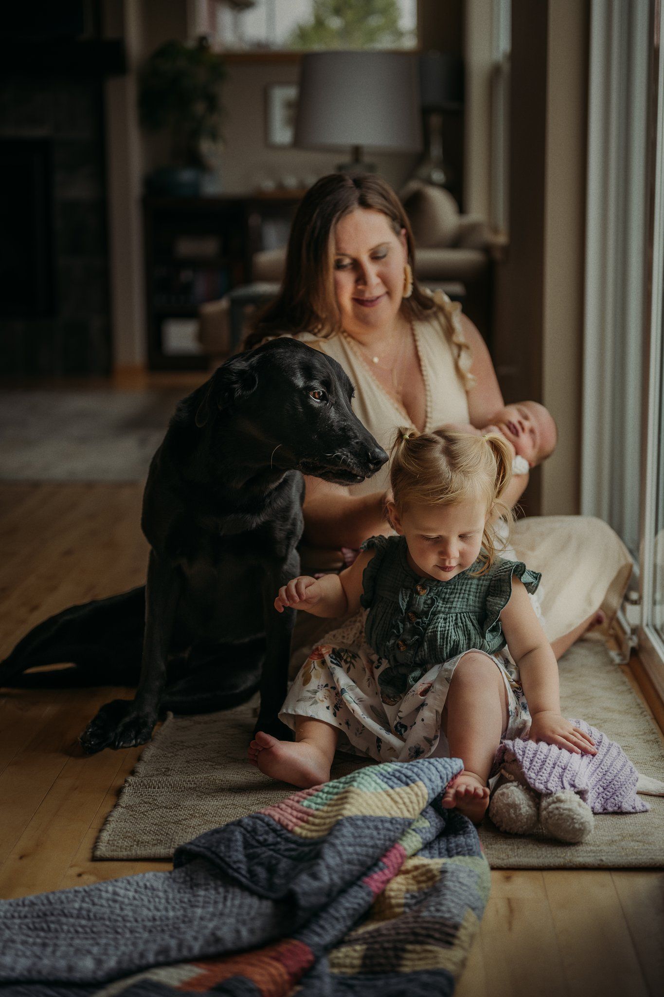 Joyful indoor family scene with toddler, infant, and black dog by sunlit window.