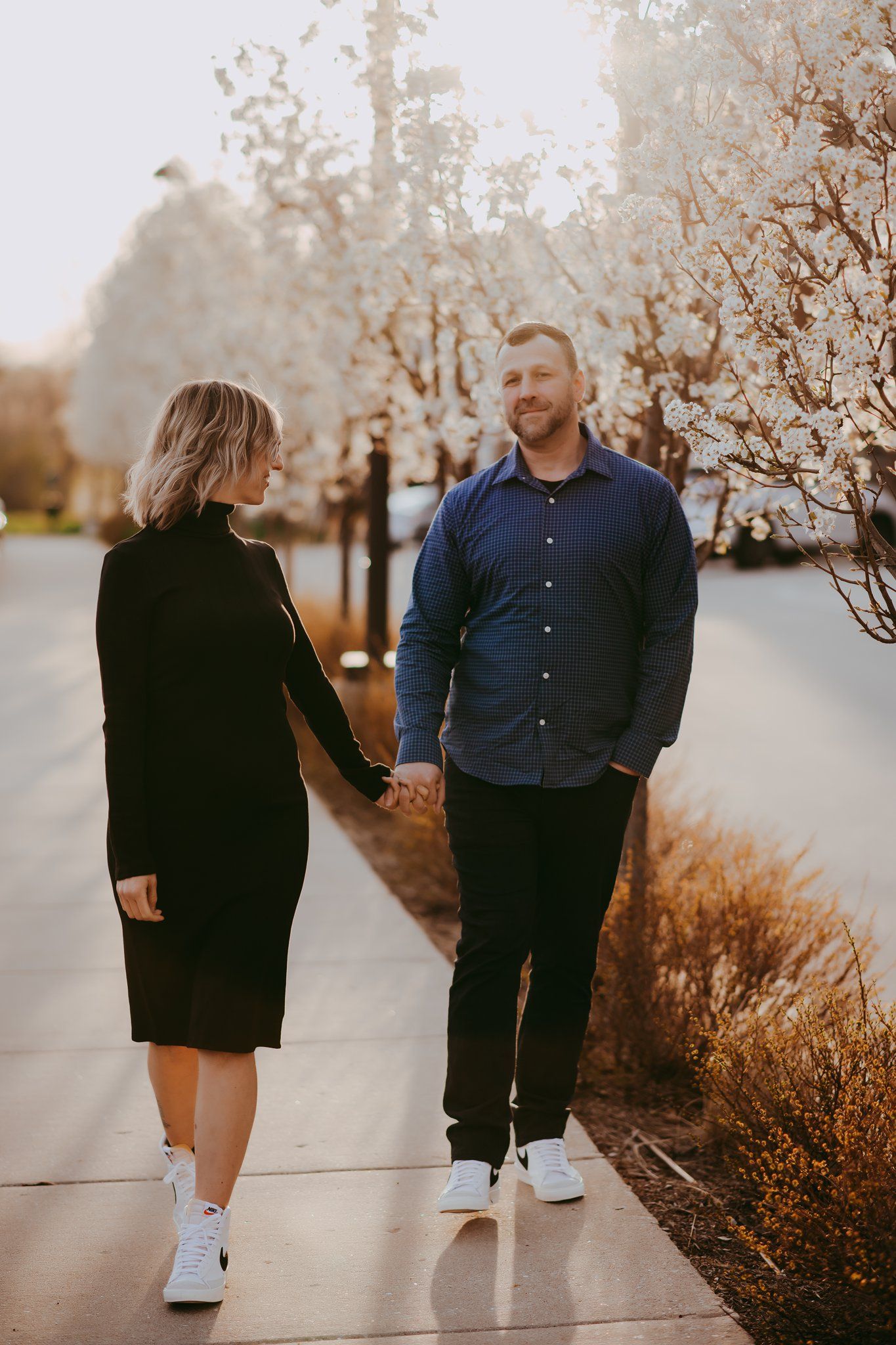 Couple holding hands while walking on sidewalk with flowering trees in background.