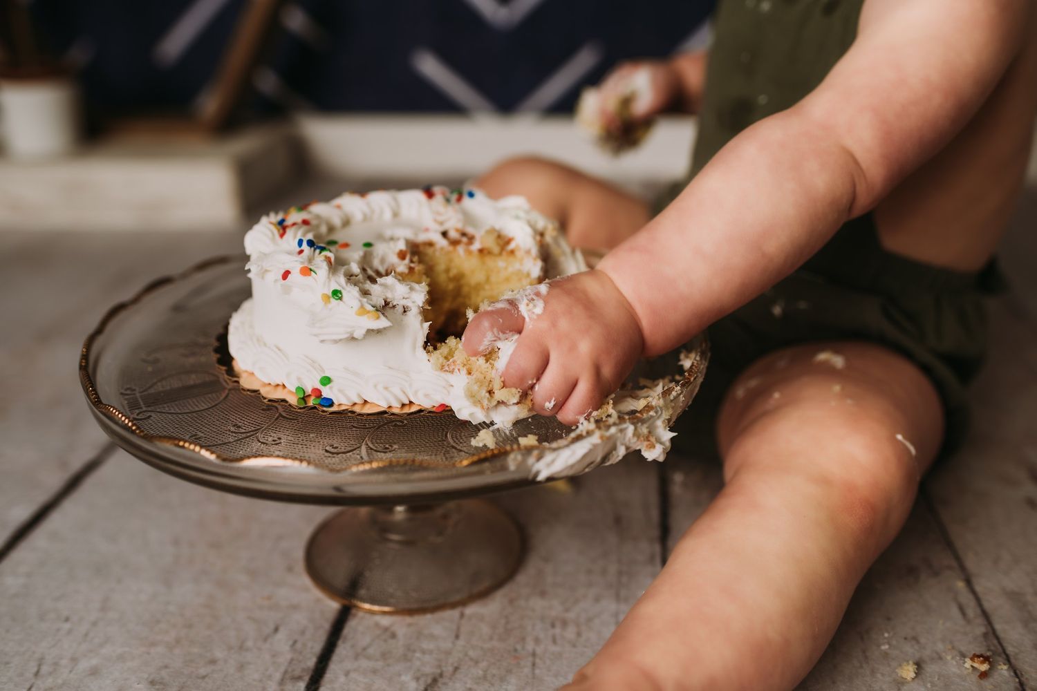 Close-up of tiny hands reaching for white frosted cake on a glass pedestal.