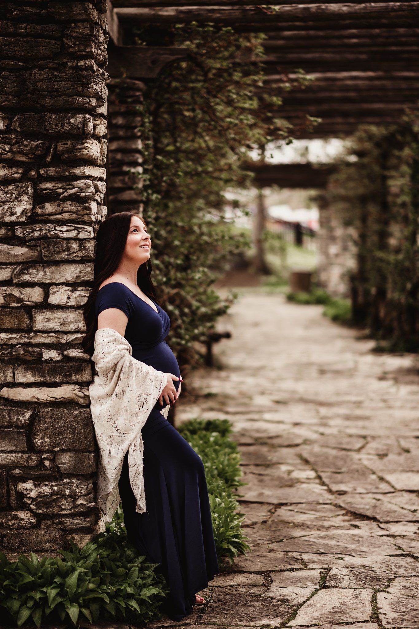 Expectant mother in navy gown poses against stone pillar under garden archway.