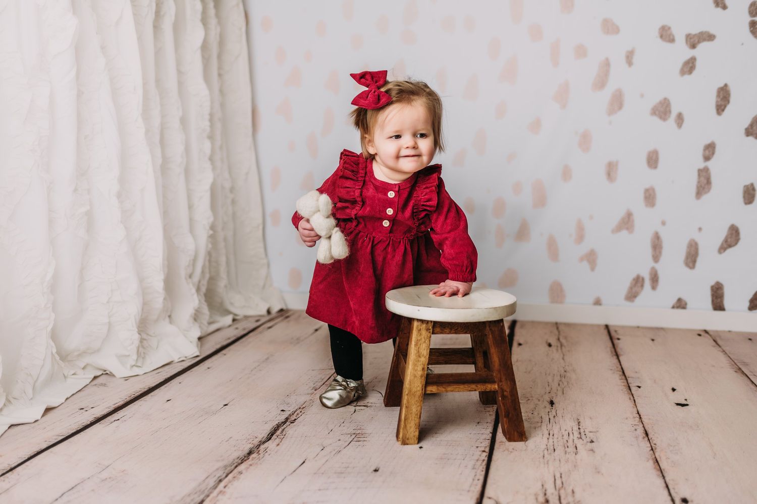 Little one in red holiday dress stands beside wooden stool against white backdrop.