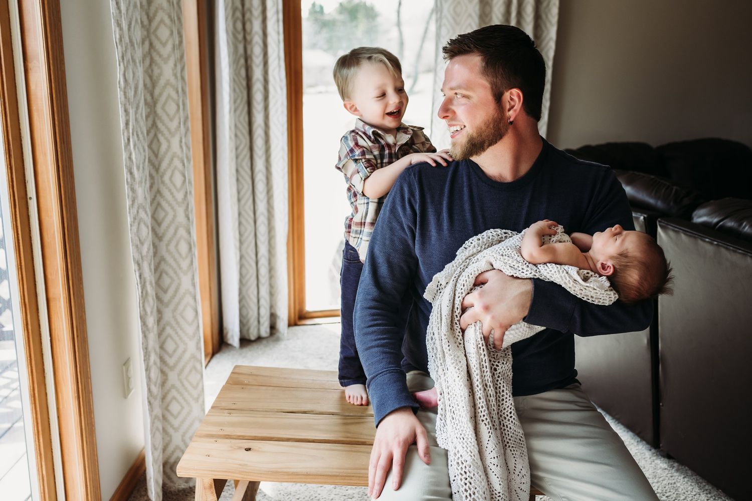 Parent balances holding two children while standing next to a wooden bench by bright windows.