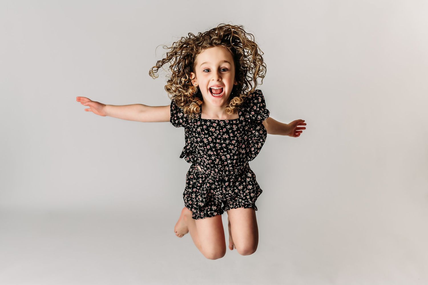An energetic child jumps mid-air wearing a black patterned romper against a white background.