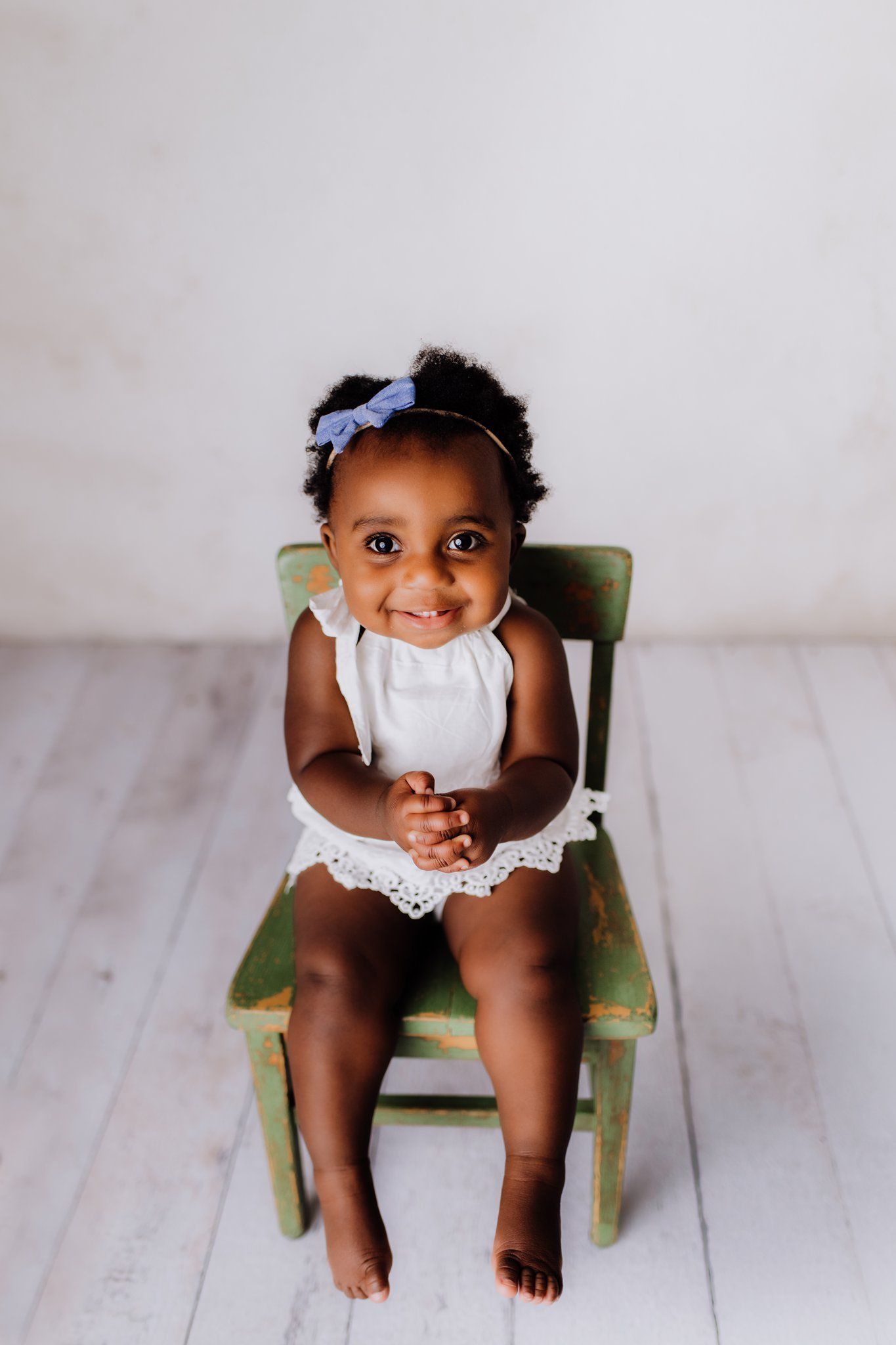 A toddler in a white dress sits on a green vintage chair against a light background.