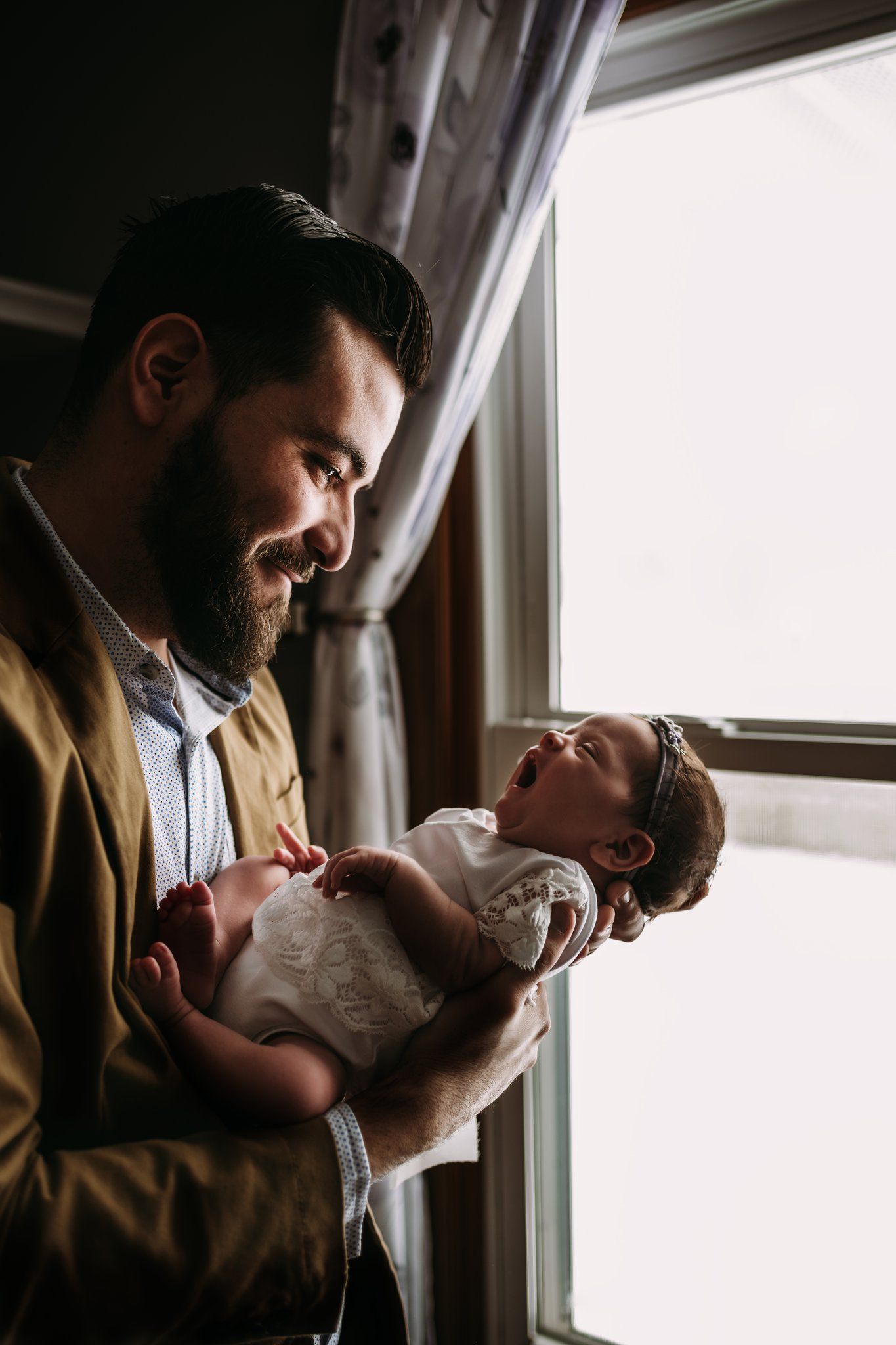 A bearded man in a tan jacket holds a small baby by a bright window.