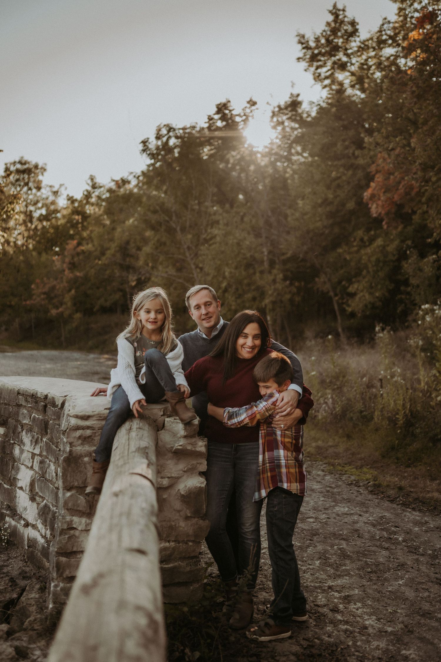 Family poses together against wooden fence railing during golden hour in natural outdoor setting.