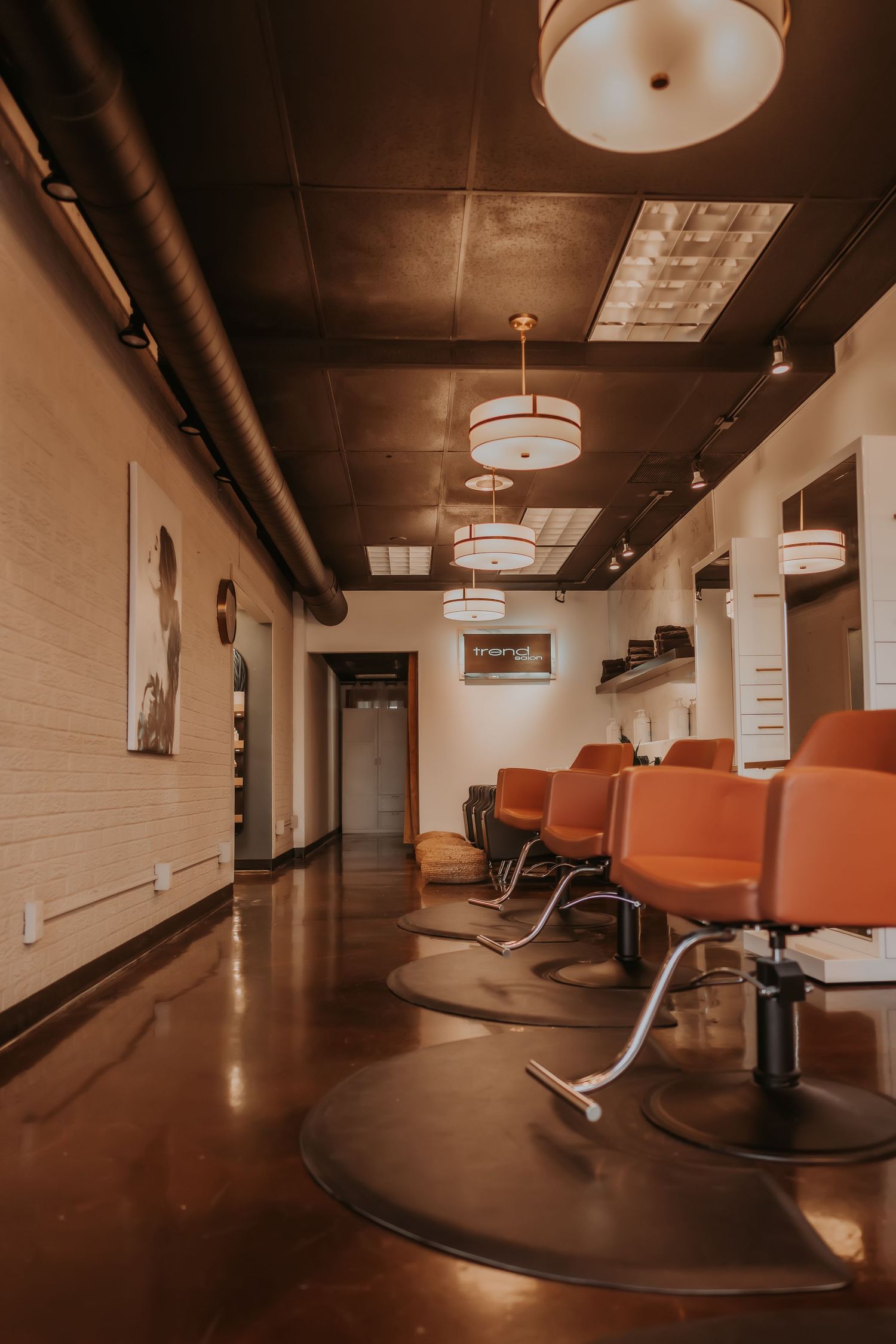 Salon corridor with polished concrete floors, orange styling chairs, and modern drum pendant lighting fixtures.
