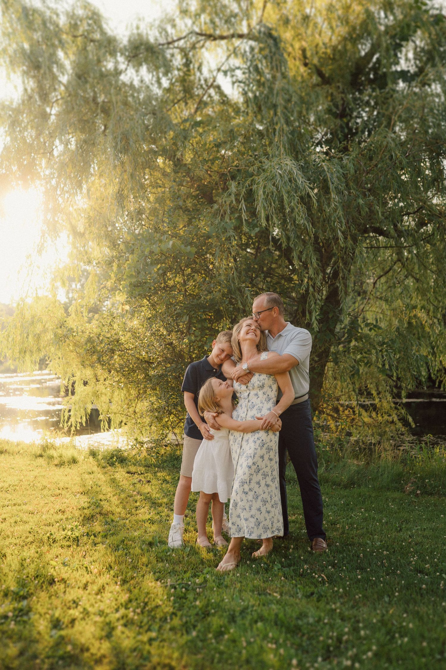 Intimate family moment captured in a sunlit garden with willow trees in the background.