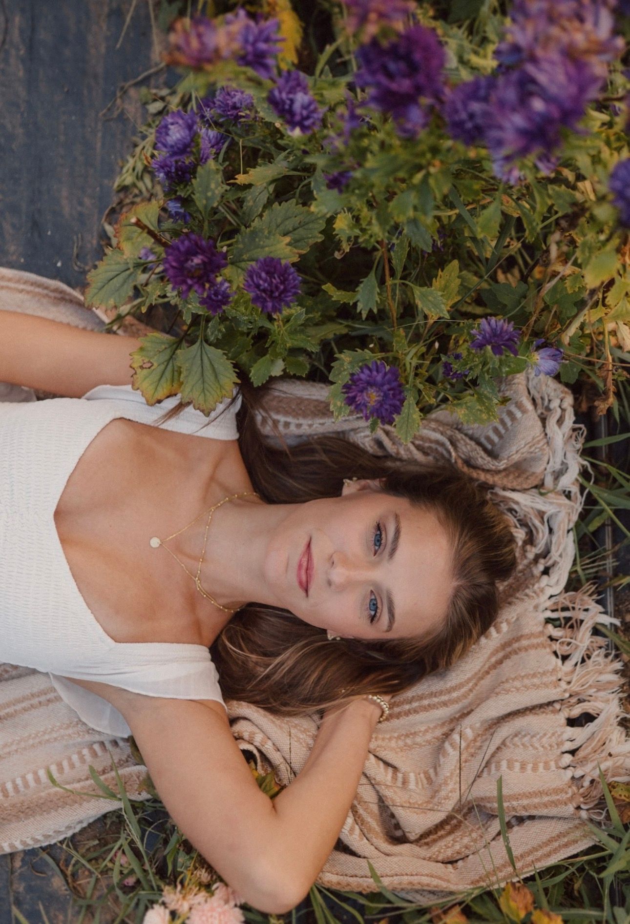 Portrait of someone lying on a blanket surrounded by purple flowers in a garden setting.