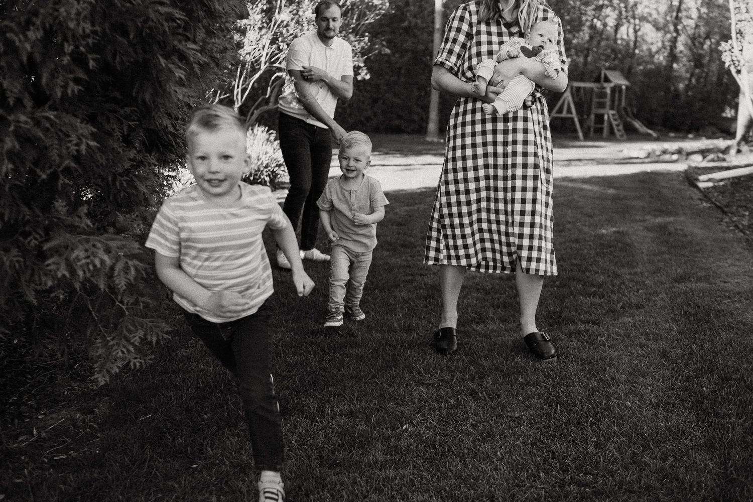 Black and white photo of children running and playing in a backyard while adults hold a baby nearby.