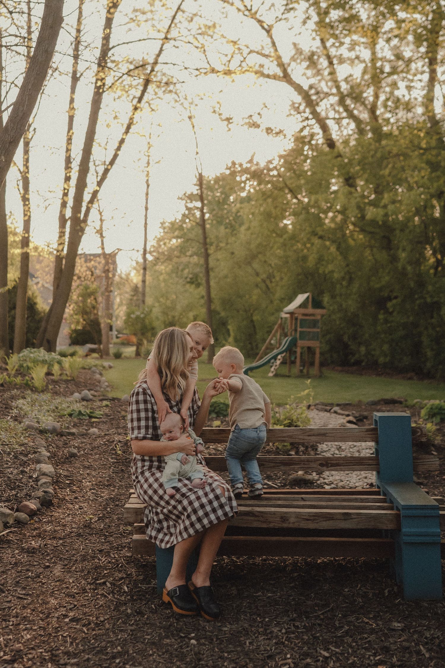 Mother sitting on garden bench with two young children in sunny backyard.