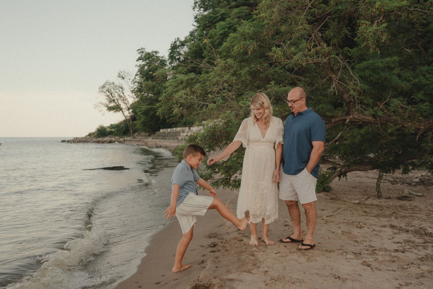 Family of three enjoying a beach walk at sunset, with a boy playfully kicking sand near the shoreline.