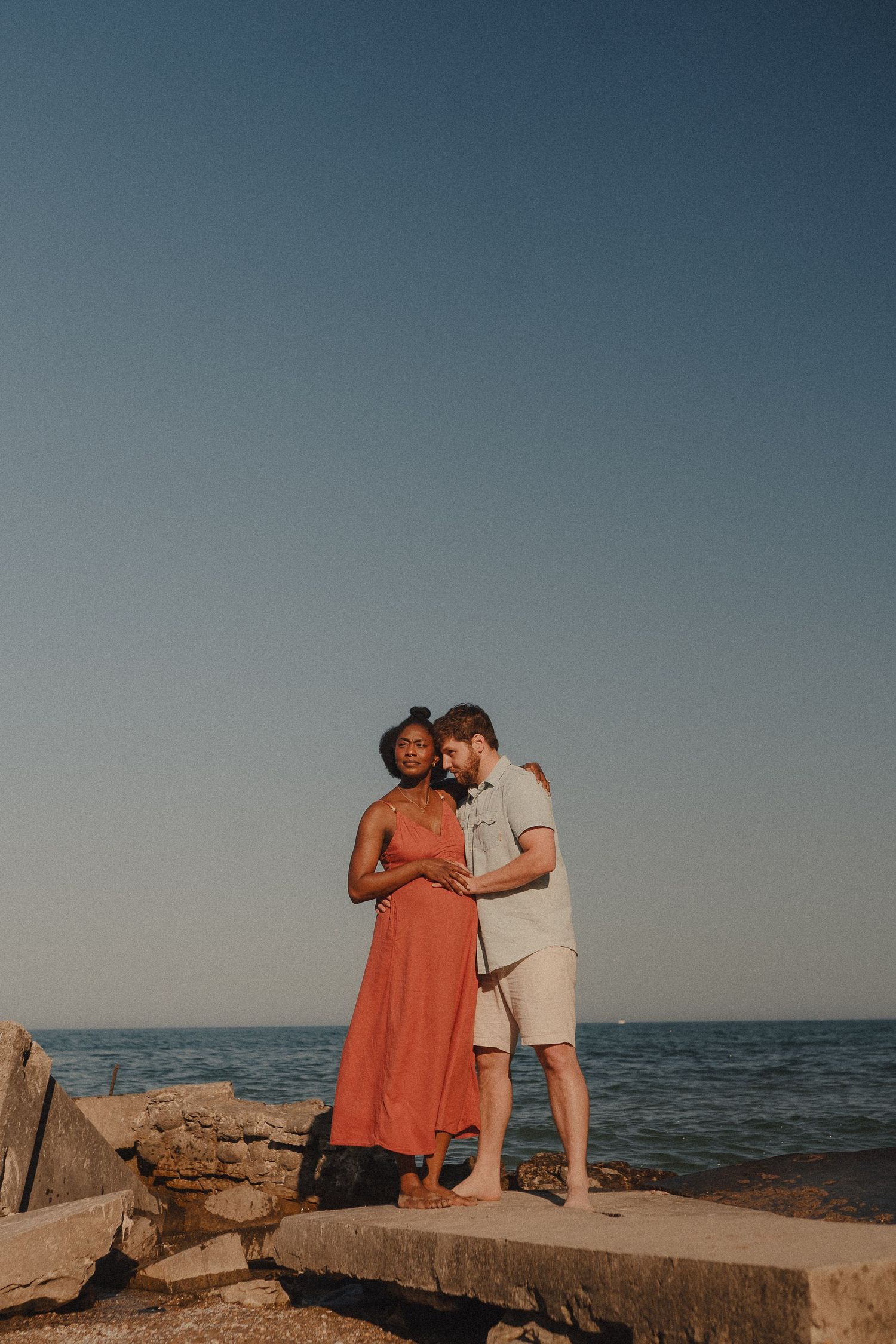 An interracial couple tenderly embraces on a rocky shoreline at sunset with calm ocean behind them.