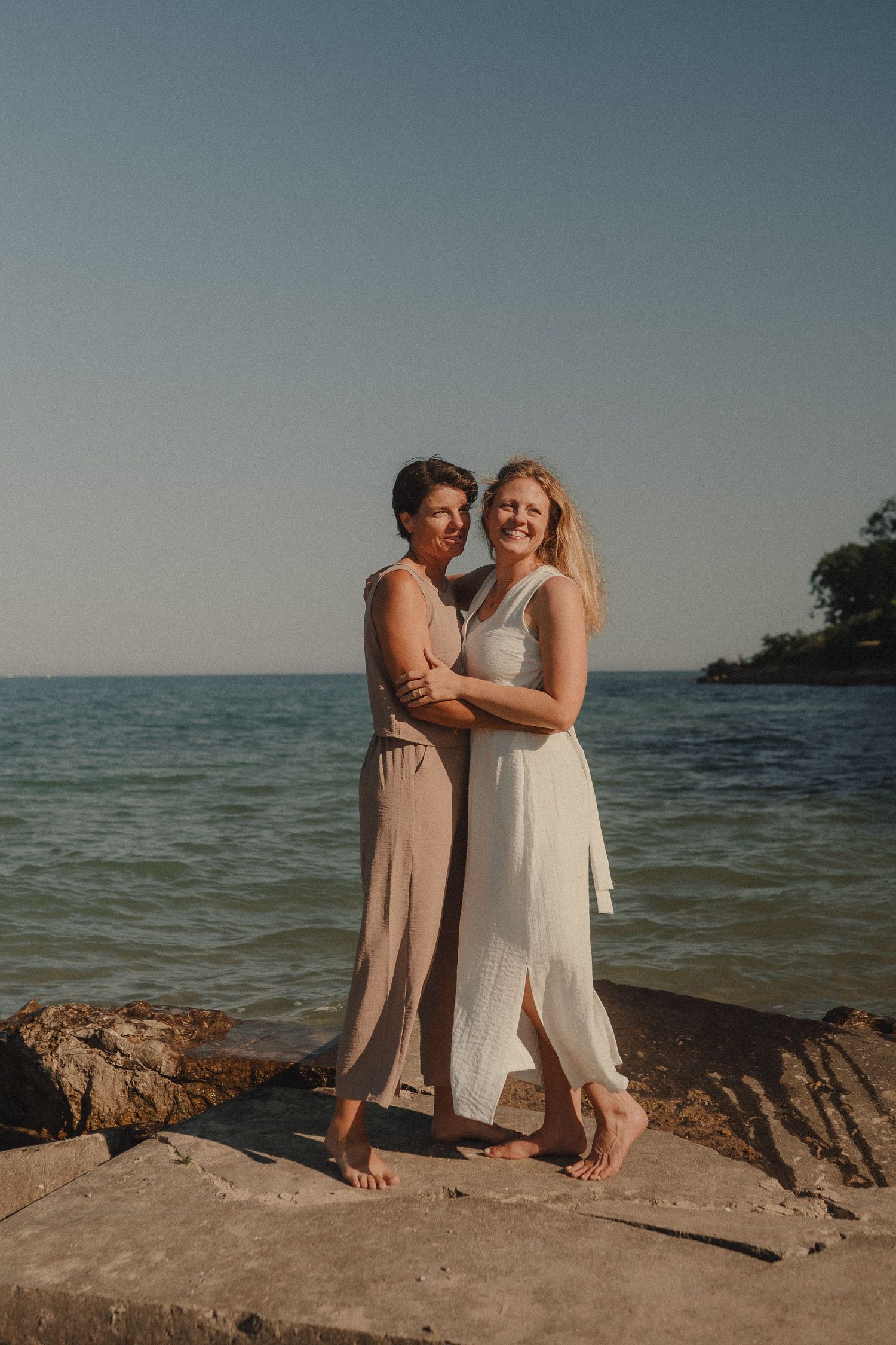 Two women embrace barefoot on rocky shoreline, wearing elegant neutral dresses with calm ocean water behind them.