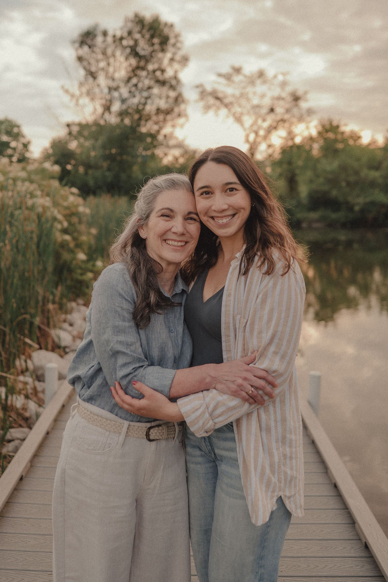 Mother and daughter embracing on a wooden dock at sunset near a peaceful pond.
