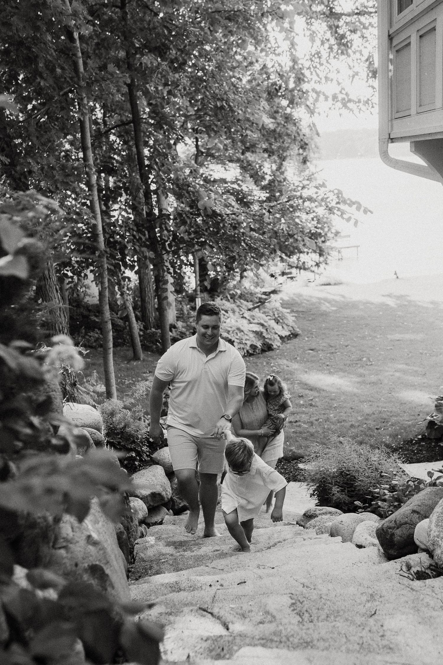 Black and white photo of father guiding young child up stone steps near lakeside home.