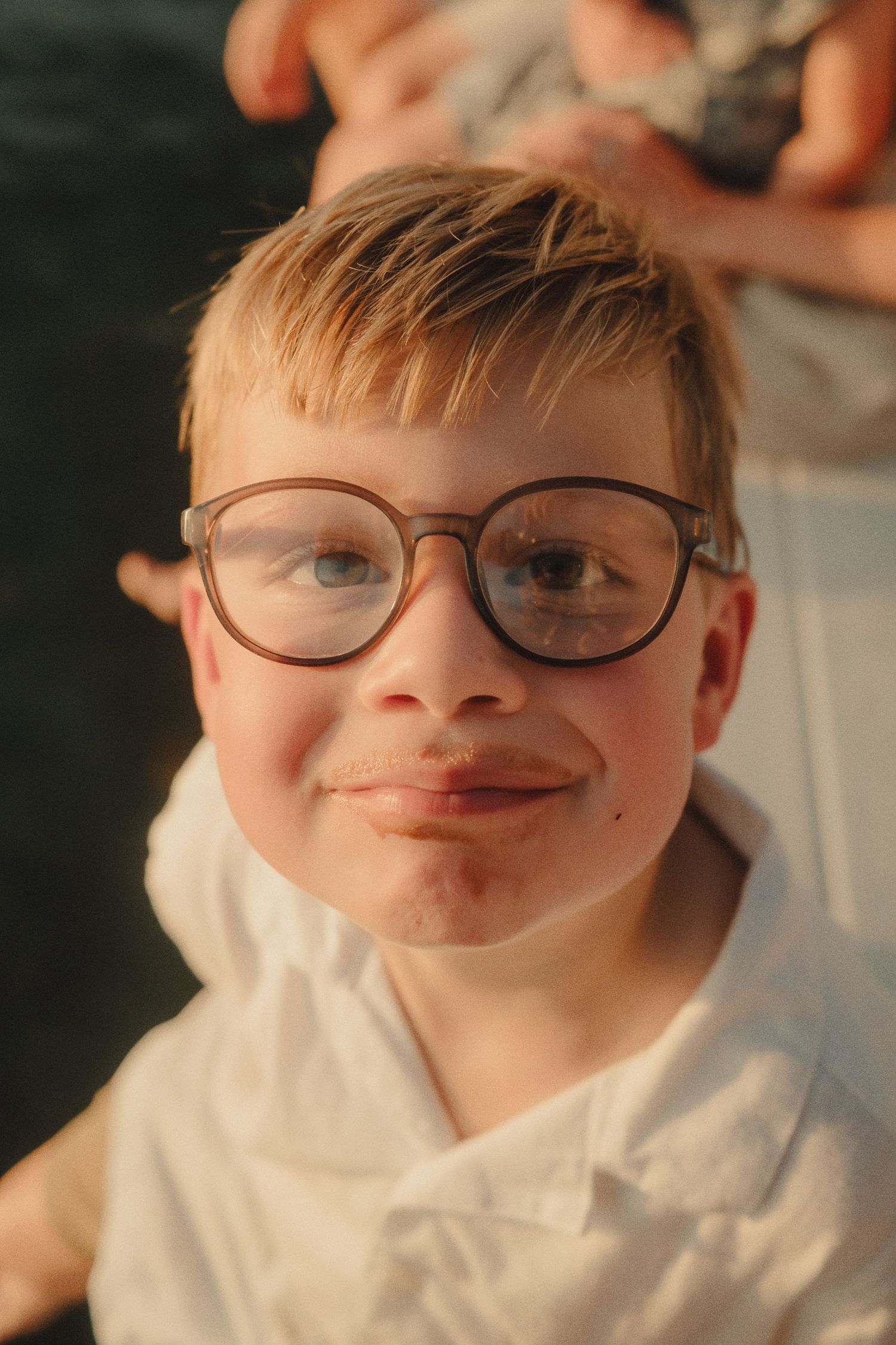 Smiling young boy with round glasses and blonde hair poses outdoors in warm golden light.