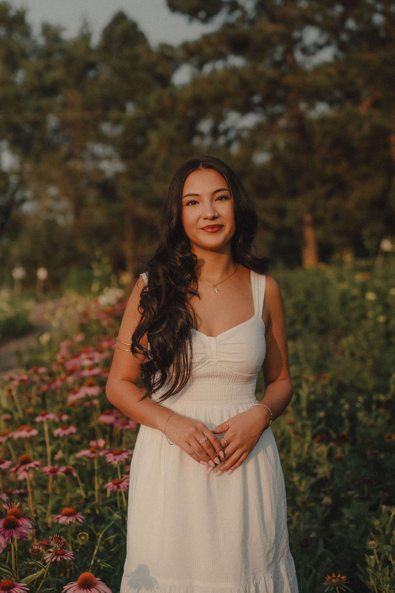 Woman in white sundress standing among pink wildflowers at golden hour with trees in background.