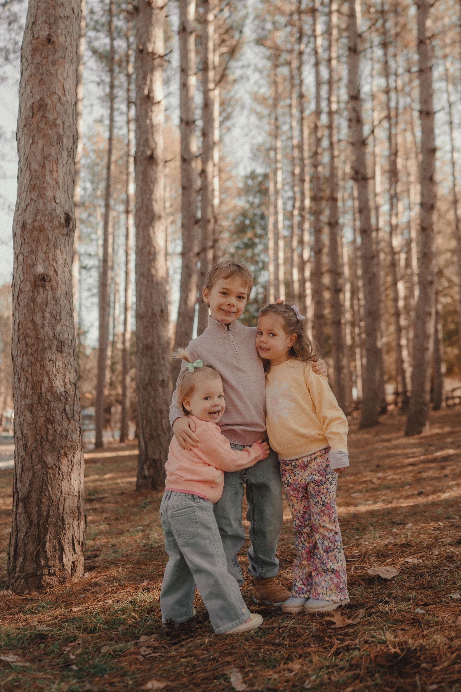Three children in cozy sweaters stand together among tall pine trees in a peaceful autumn forest setting.