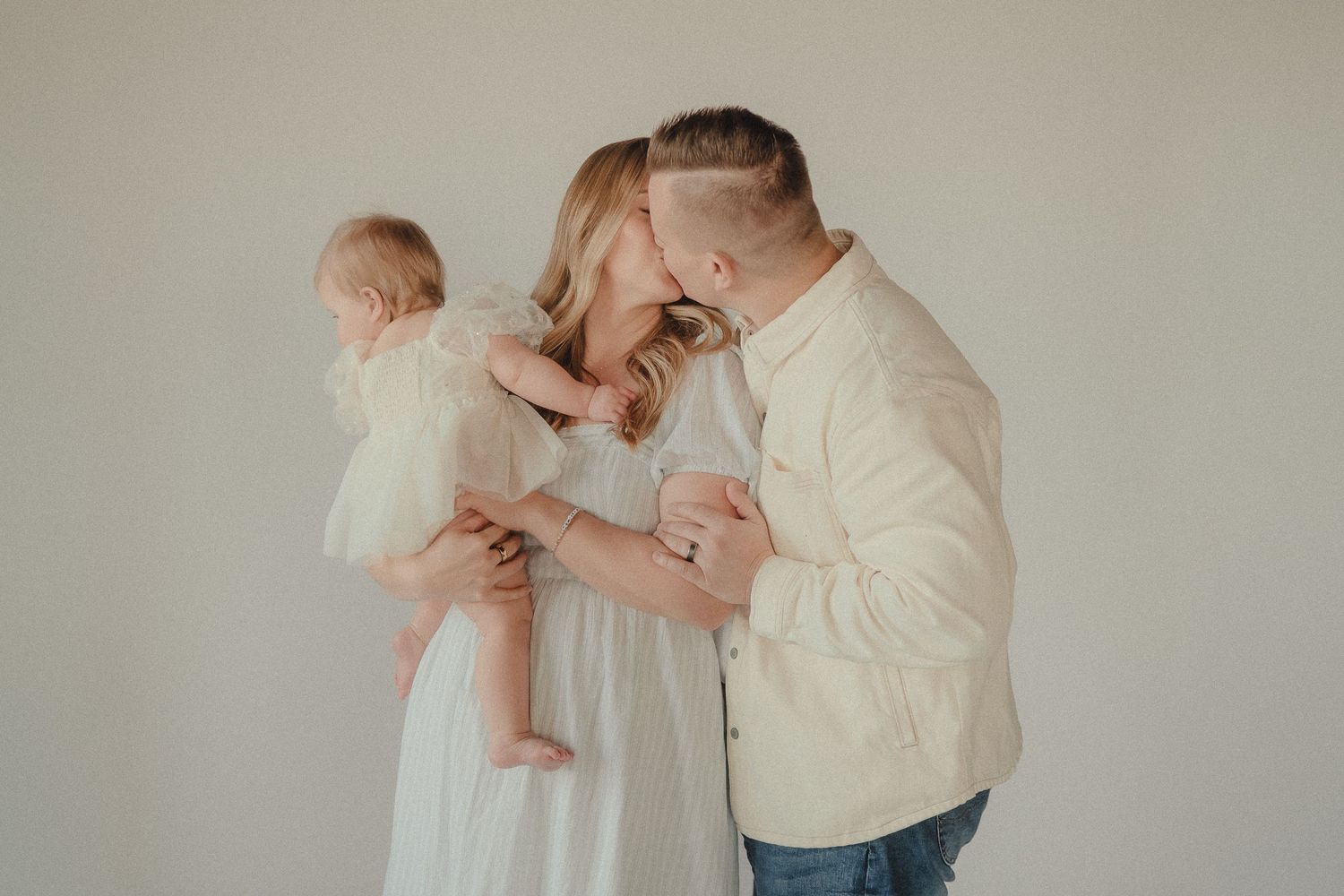 A couple shares a tender kiss while holding their baby girl dressed in white in a bright studio setting.