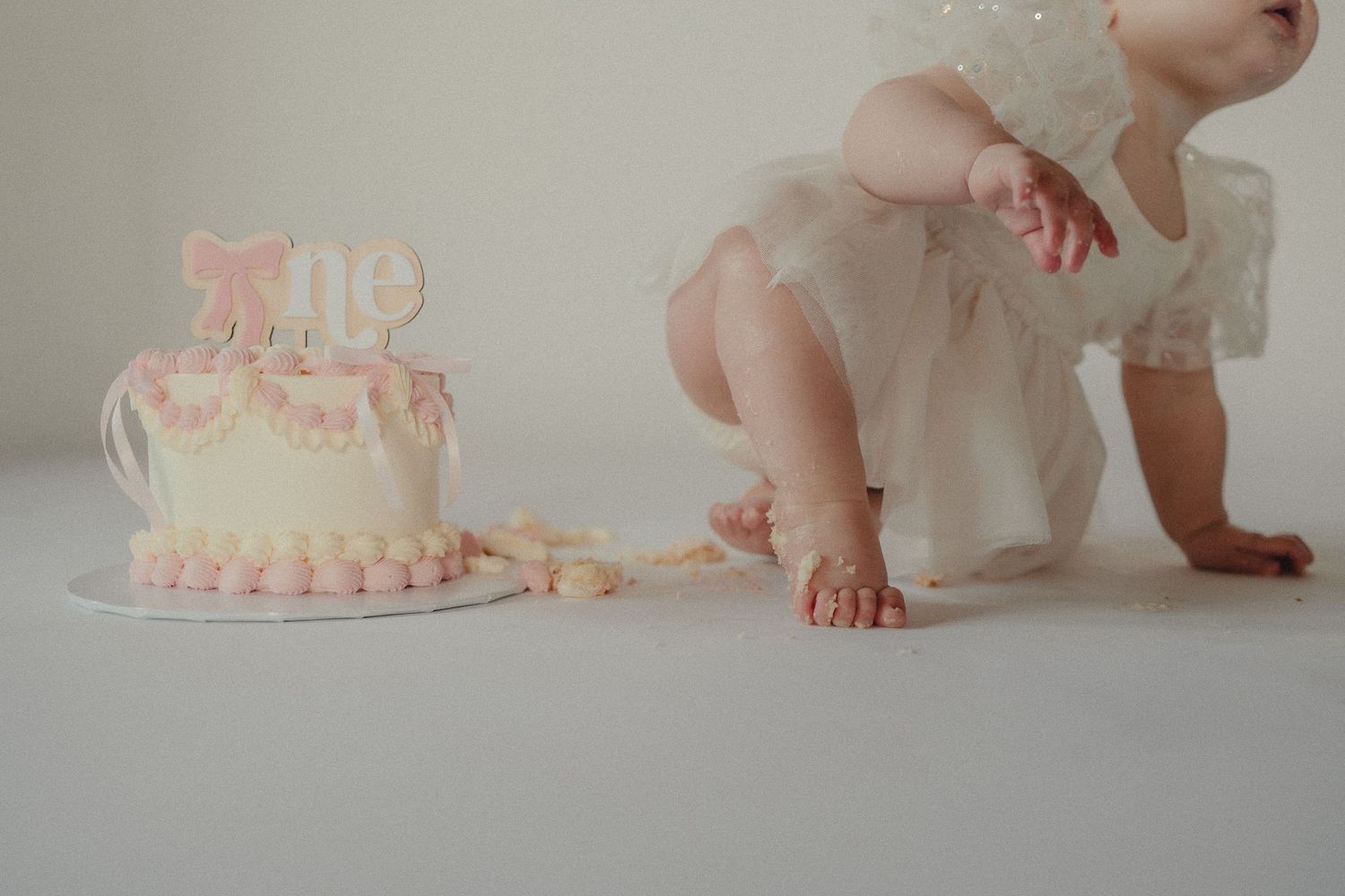 Baby girl in white dress beside a pink first birthday smash cake on a white background.