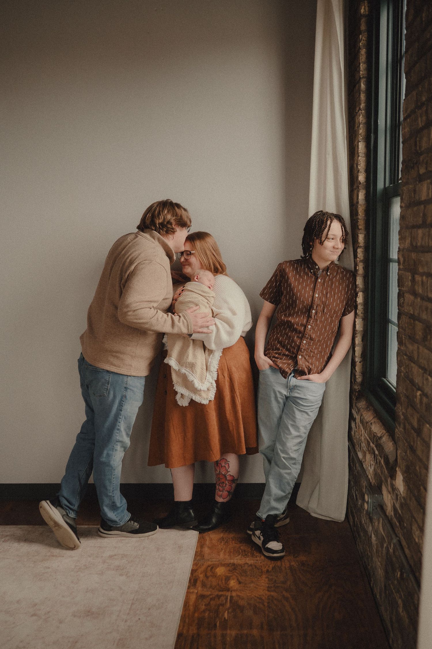 Parents lovingly kiss newborn baby while older sibling leans against wall near curtained window.